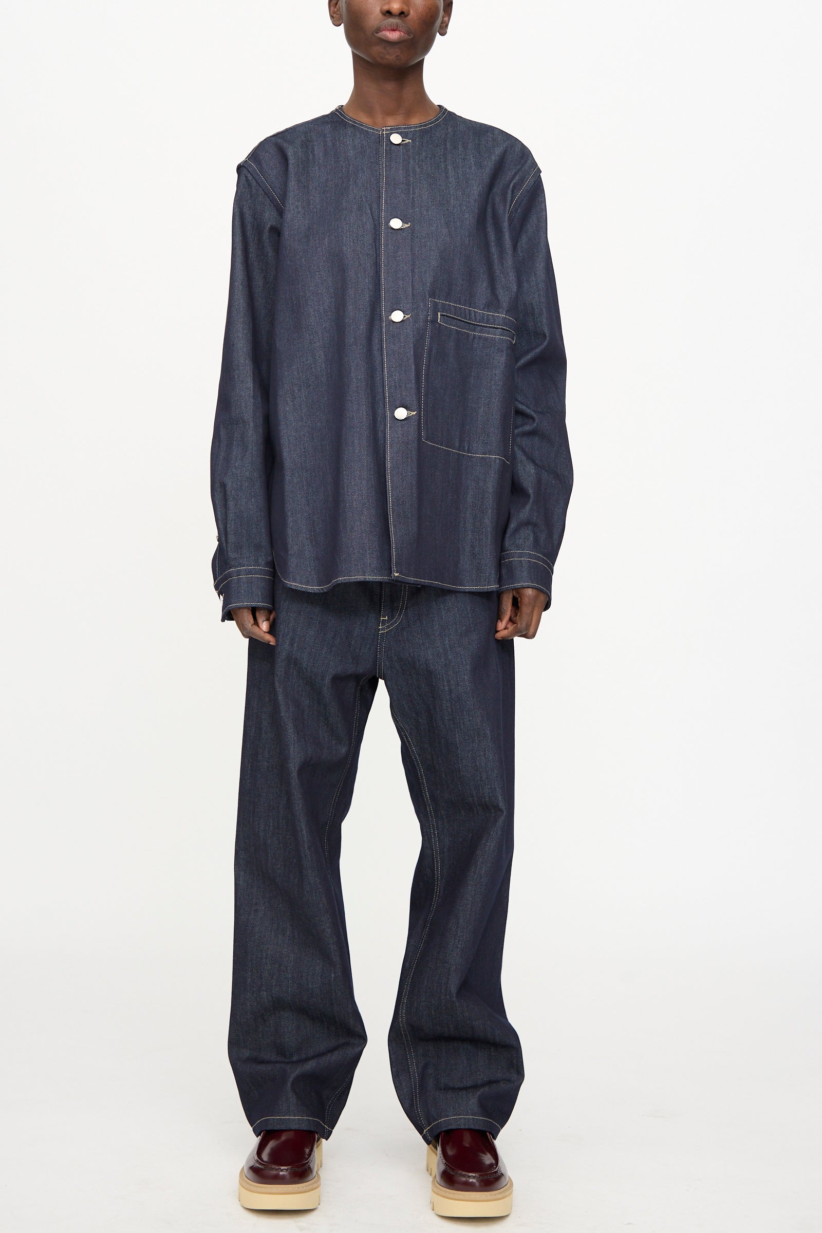 A person wearing the Sofie D'Hoore Barna Shirt in Midnight—a relaxed fit, long sleeve raw denim top with a front pocket—paired with matching wide-leg denim pants and burgundy platform shoes, stands against a plain white background.