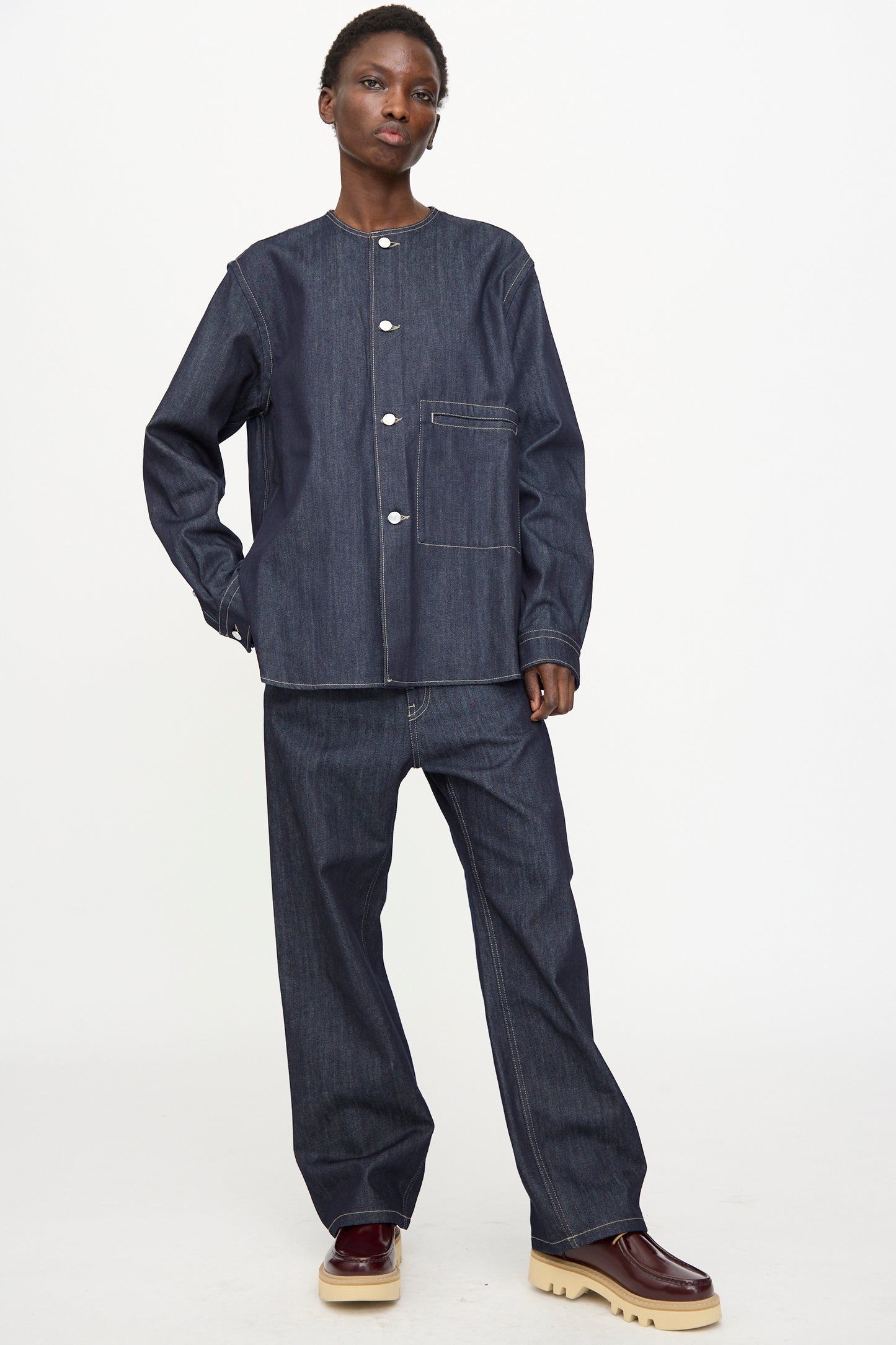 A person stands in a relaxed pose wearing the Sofie D'Hoore Barna Shirt in Midnight with matching pants and beige-soled burgundy shoes against a plain white background.