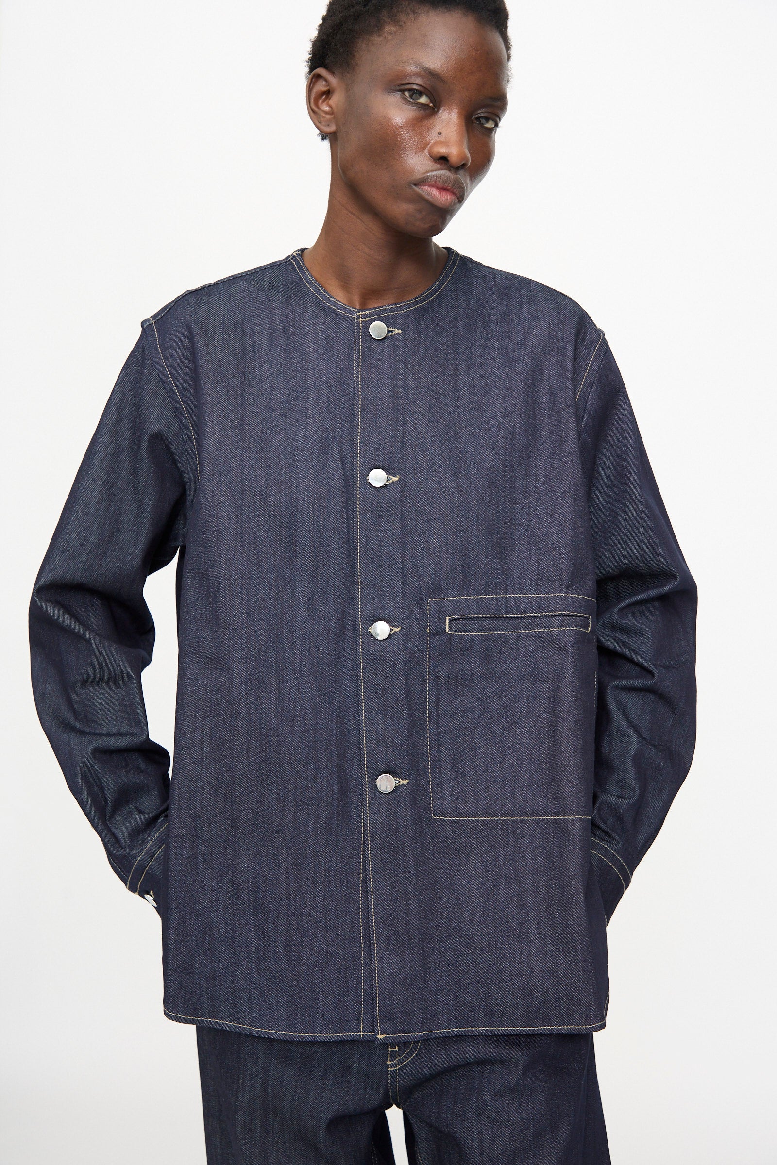 A person wears the Sofie D'Hoore Barna Shirt in Midnight, a relaxed-fit dark denim long-sleeve jacket with a round collar, white stitching, front buttons, and a chest pocket. Matching denim pants complete the look.