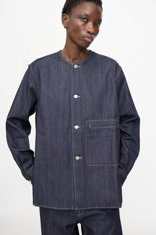 A person wears the Sofie D'Hoore Barna Shirt in Midnight, a relaxed-fit dark denim long-sleeve jacket with a round collar, white stitching, front buttons, and a chest pocket. Matching denim pants complete the look.