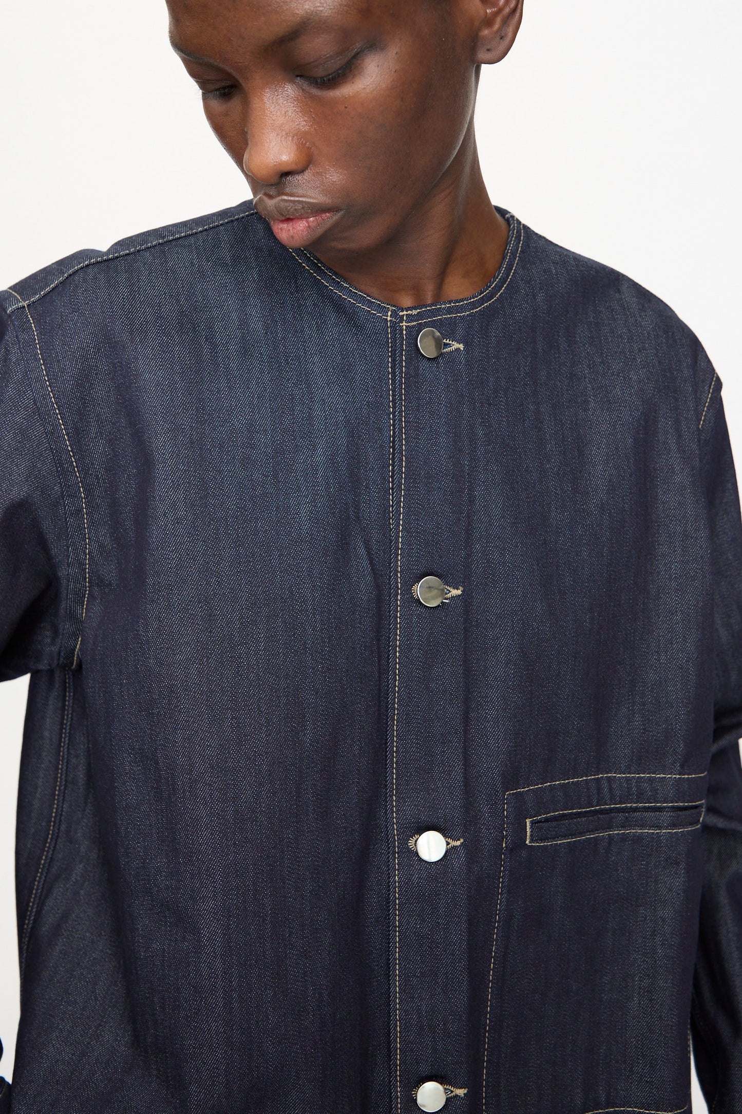 A person wearing the Sofie D'Hoore Barna Shirt in Midnight—a relaxed fit dark denim jacket with white stitching, silver buttons, and a chest pocket—looks downward.