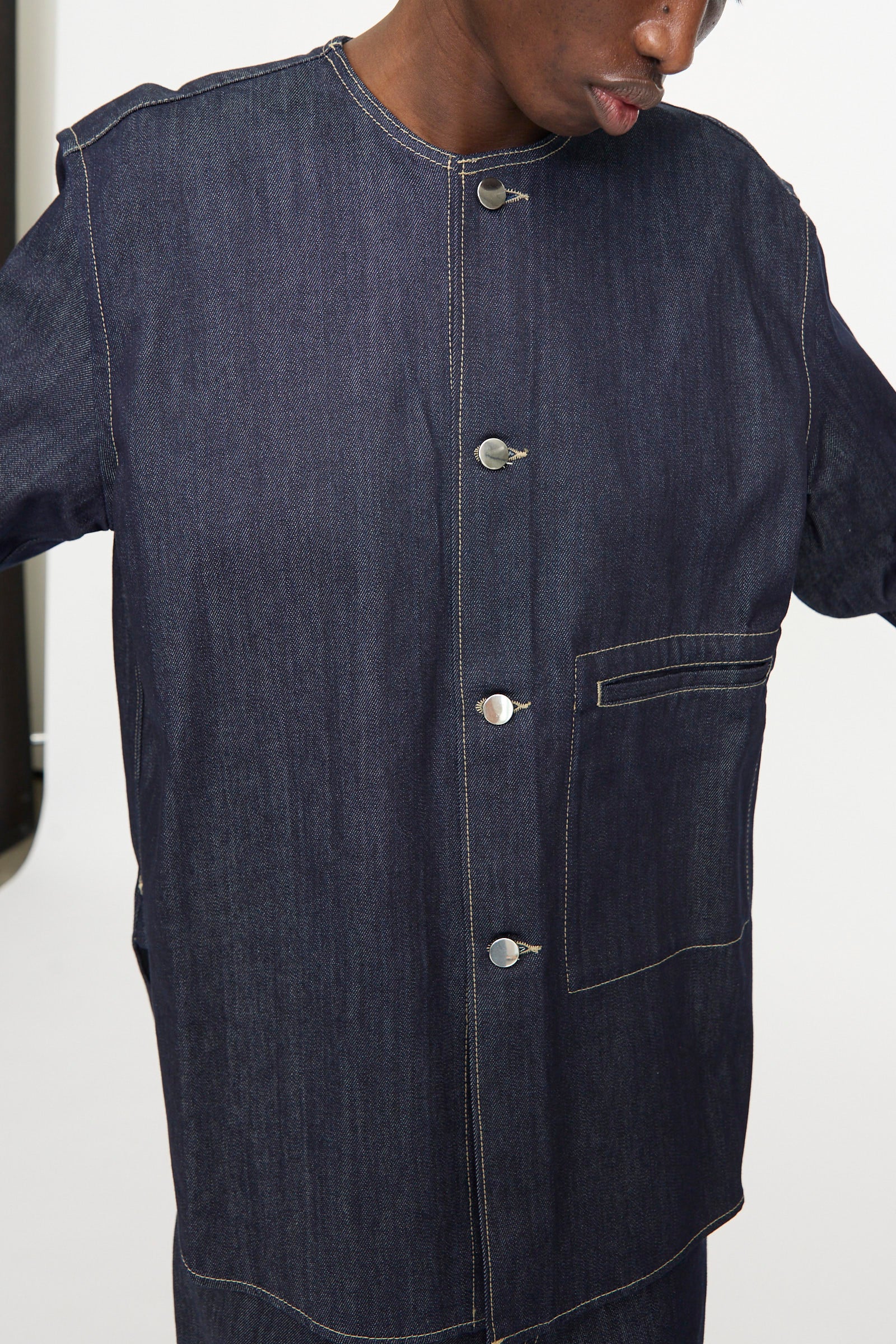 A person shown from shoulders to waist wears the Sofie D'Hoore Barna Shirt in Midnight, featuring a relaxed fit, dark denim fabric, metal buttons, and a large front pocket.