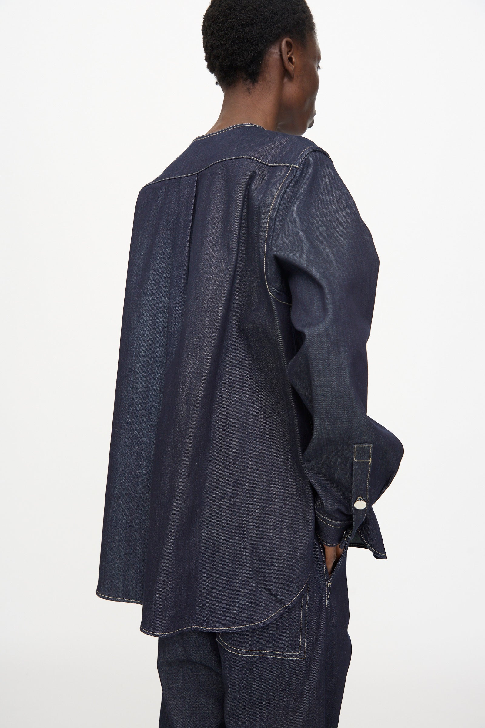 A person stands sideways against a plain white background wearing the Sofie D'Hoore Barna Shirt in Midnight, a relaxed fit long sleeve shirt in dark denim with visible stitching, paired with matching denim pants.