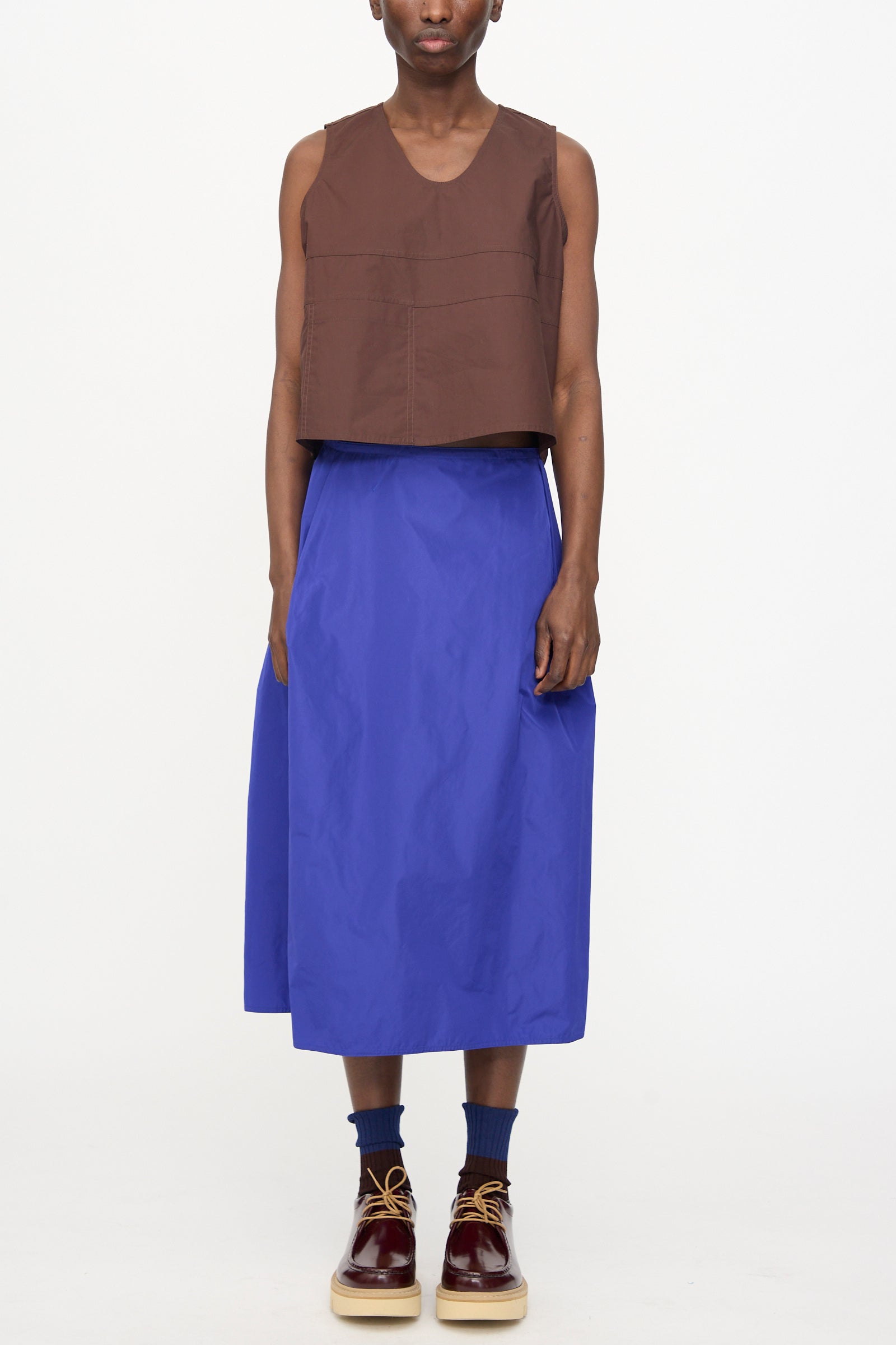 A person wears the Sofie D'Hoore Brambel Tank Shirt in Cacao, a blue midi skirt, blue socks, and brown lace-up shoes, standing against a plain white background.