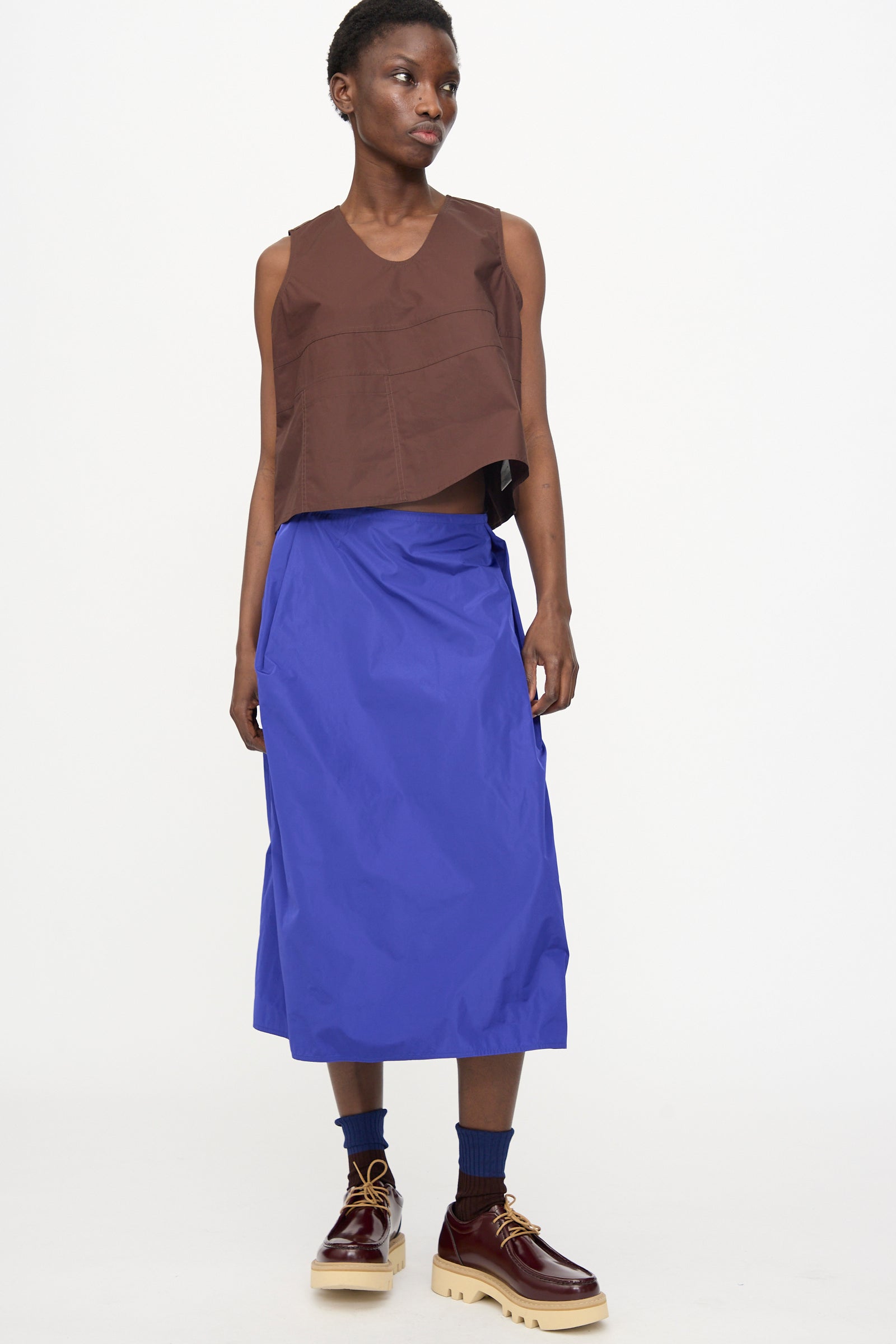 Model wears the Brambel Tank Shirt in Cacao by Sofie D'Hoore with a blue midi skirt, blue socks, and brown platform shoes, standing against a plain white background.