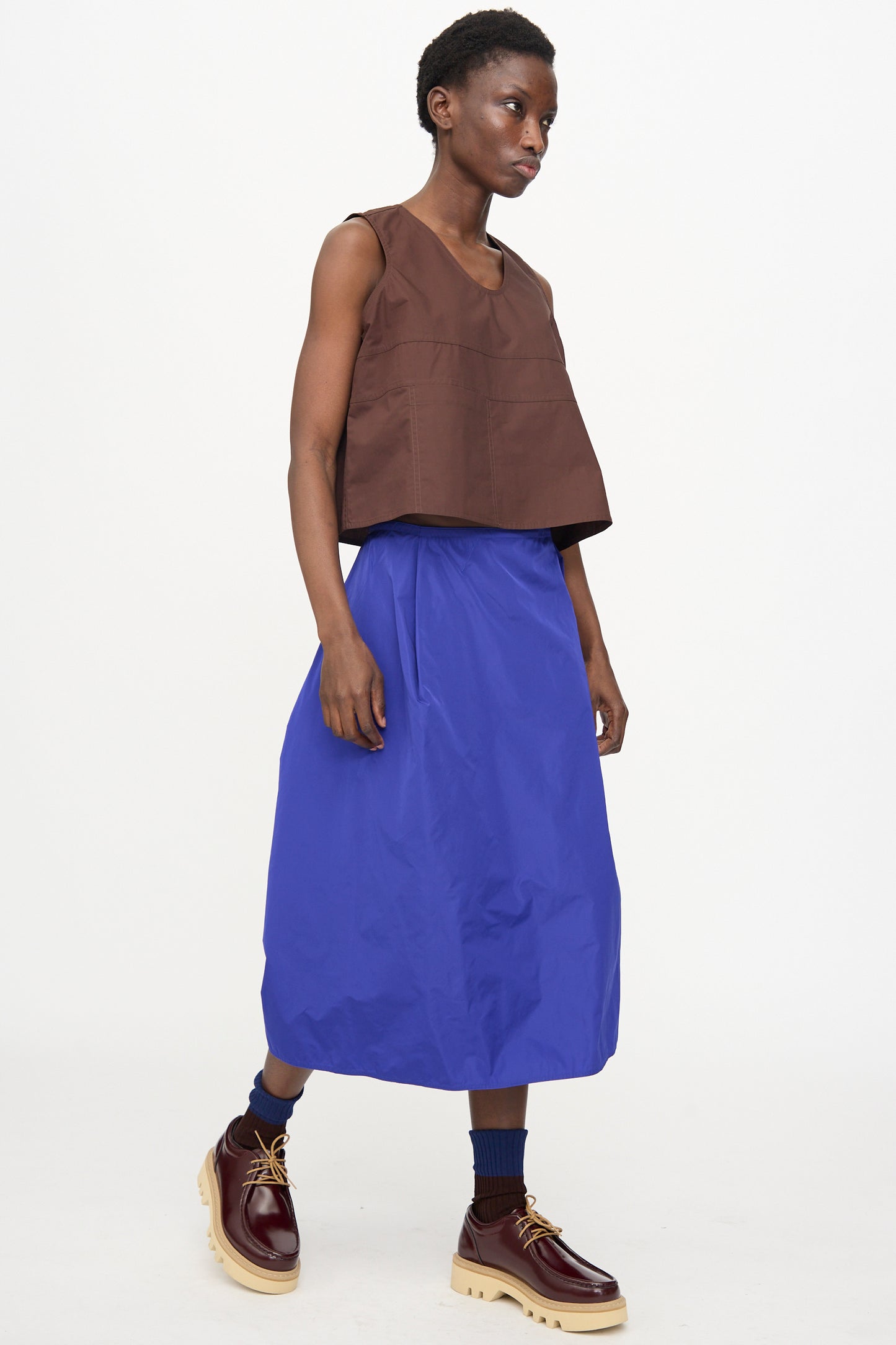 A person wearing the Brambel Tank Shirt in Cacao by Sofie D'Hoore, a blue midi skirt, navy socks, and chunky brown shoes walks against a plain white background.