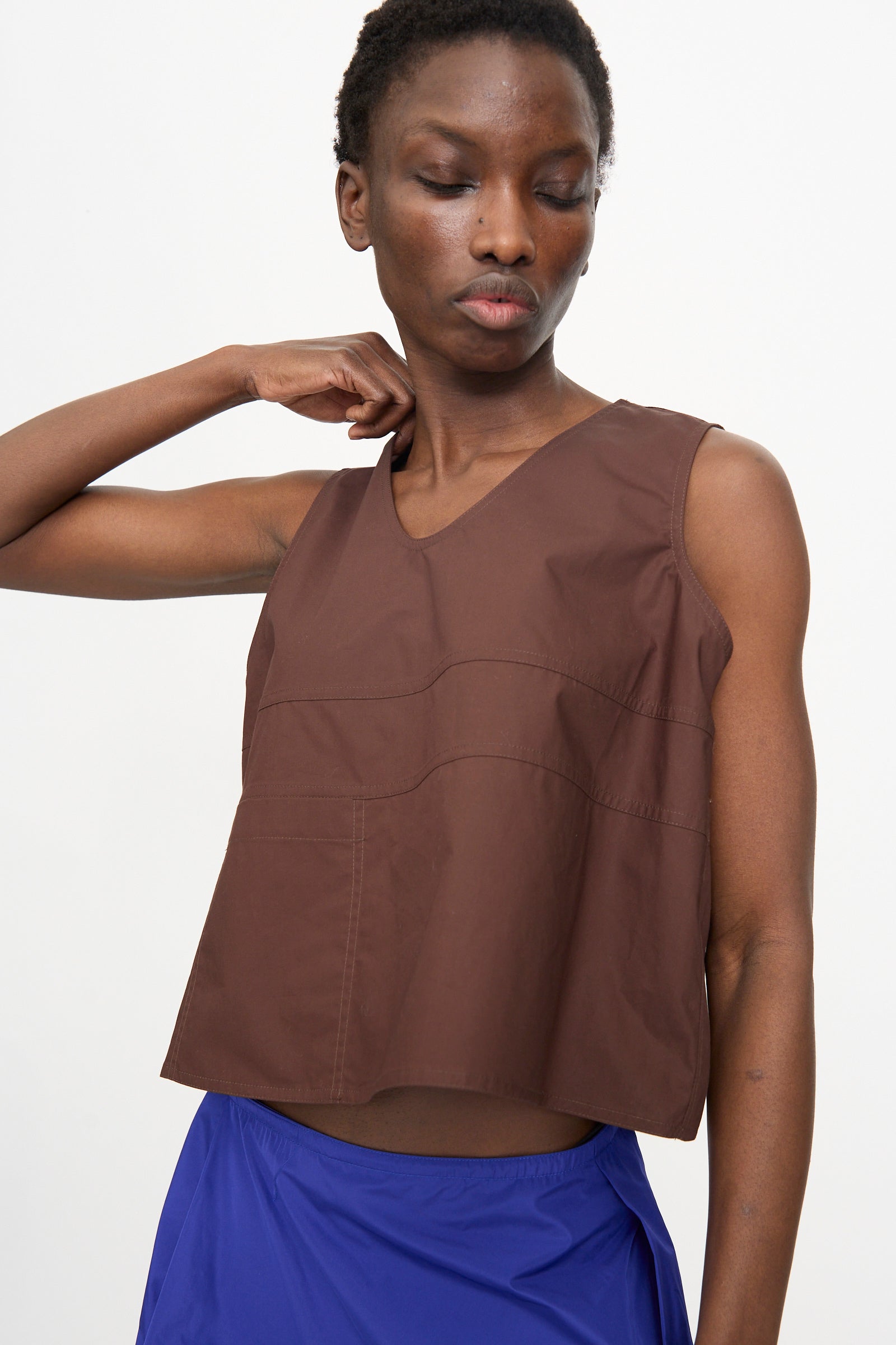 A person wears the Sofie D'Hoore Brambel Tank Shirt in Cacao, a boxy fit sleeveless brown cotton top, paired with blue bottoms and stands against a plain light background, looking down with one hand raised to their neck.