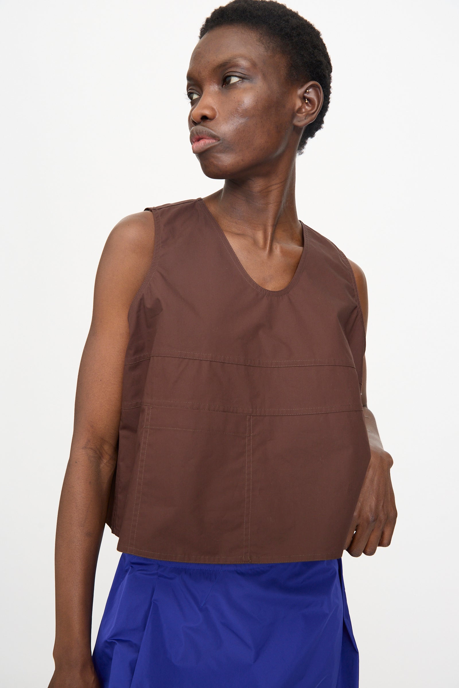 A person wears the Sofie D'Hoore Brambel Tank Shirt in Cacao with blue bottoms, standing against a plain white background and looking to the side.