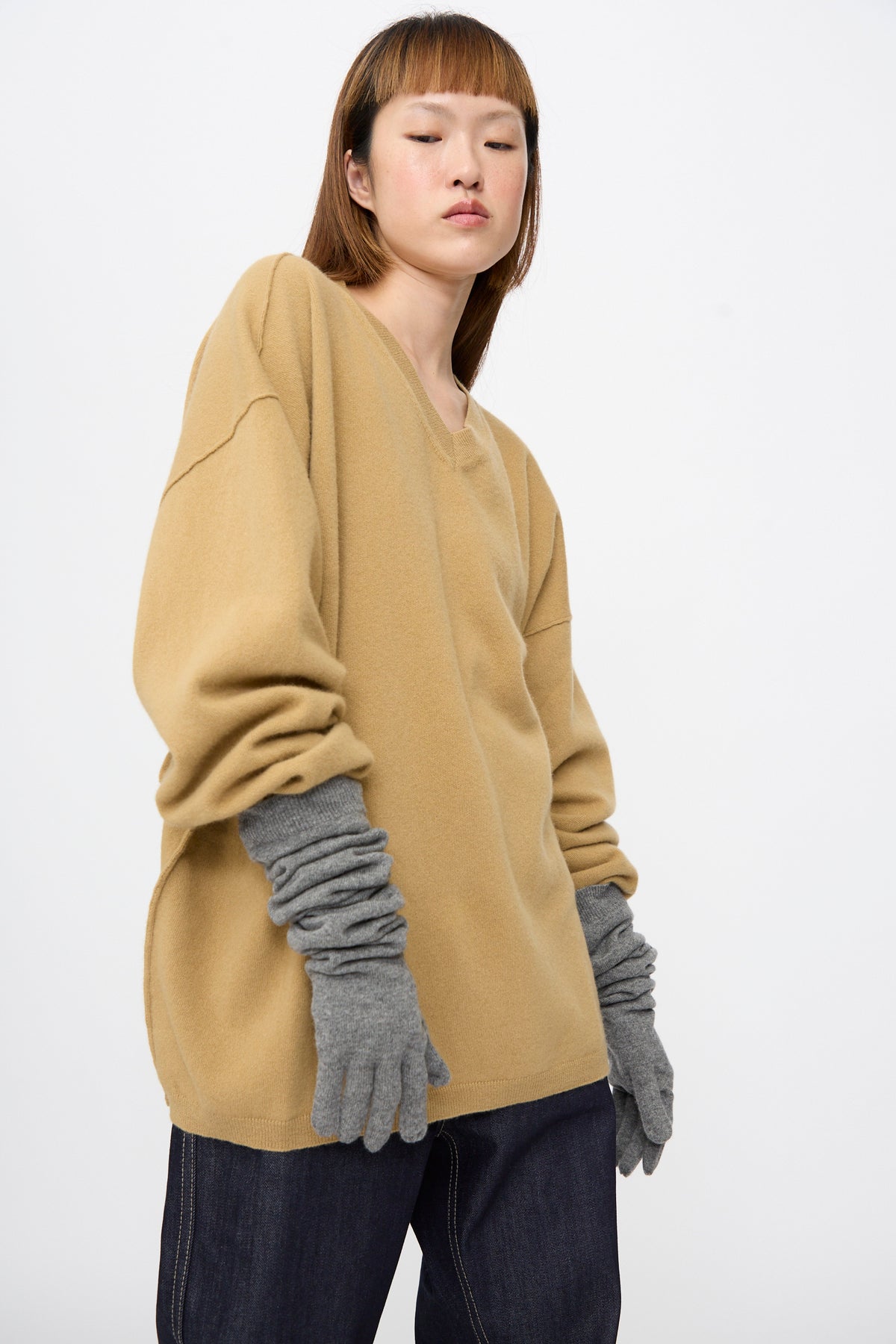 A person wearing a tan V-neck sweater and Sofie D’Hoore Cashmere Knit Alta Gloves in Metal Melange, which are long, grey, and feature ribbed cuffs.