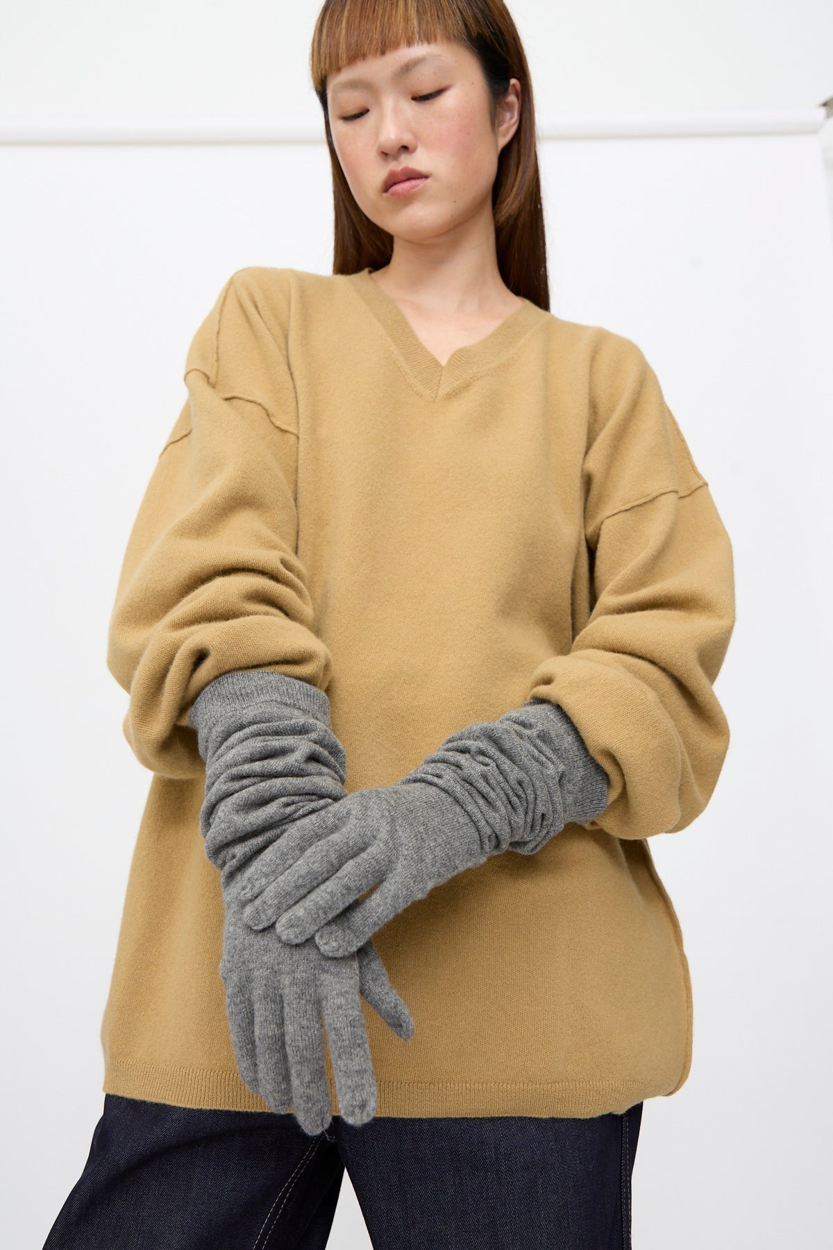 A person wearing a tan sweater and Sofie D’Hoore Cashmere Knit Alta Gloves in Metal Melange, featuring grey over-elbow length and ribbed cuffs.