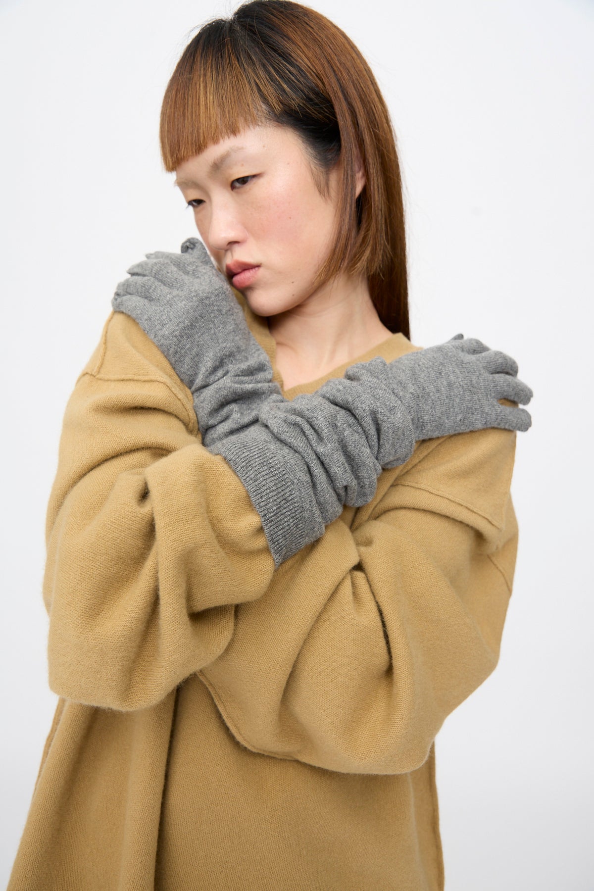 A person wearing grey Sofie D’Hoore Cashmere Knit Alta Gloves in Metal Melange, featuring ribbed cuffs that are scrunched to the mid-forearm, worn over a tan sweater.