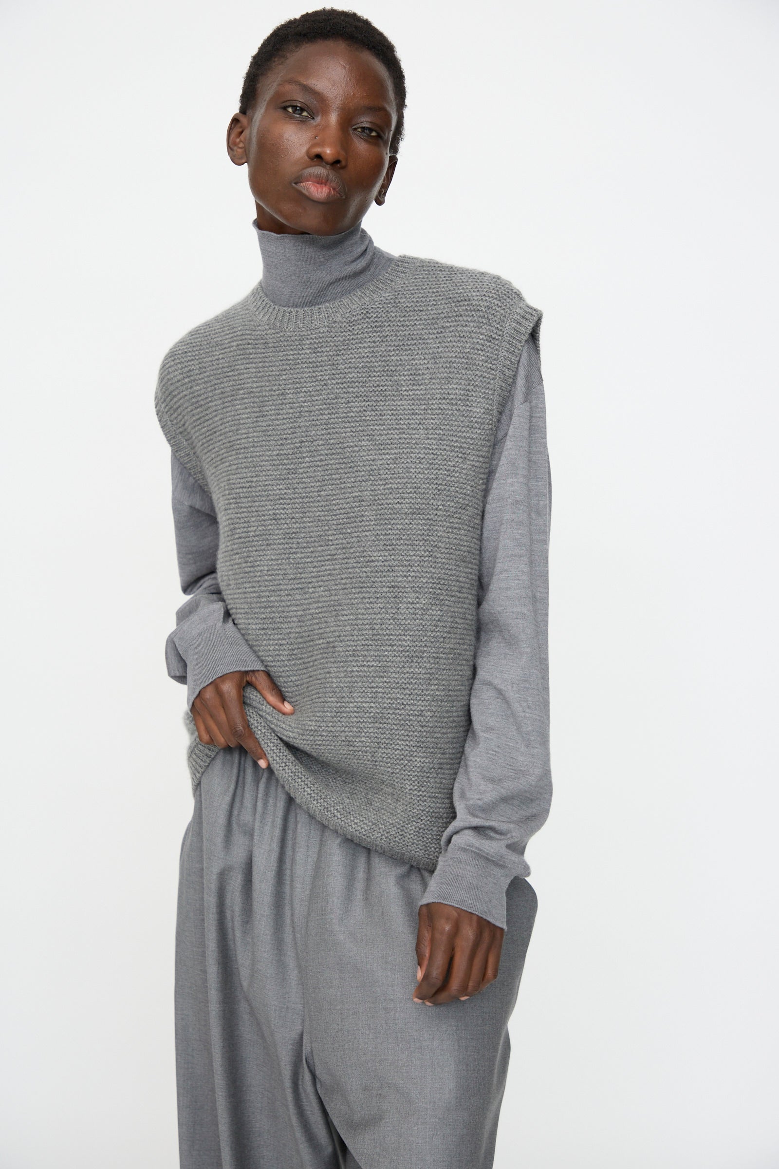 A person wears the Sofie D'Hoore Cashmere Matta Sweater in Mid Grey Melange, a sleeveless textured vest, and loose grey pants, standing against a plain white background.