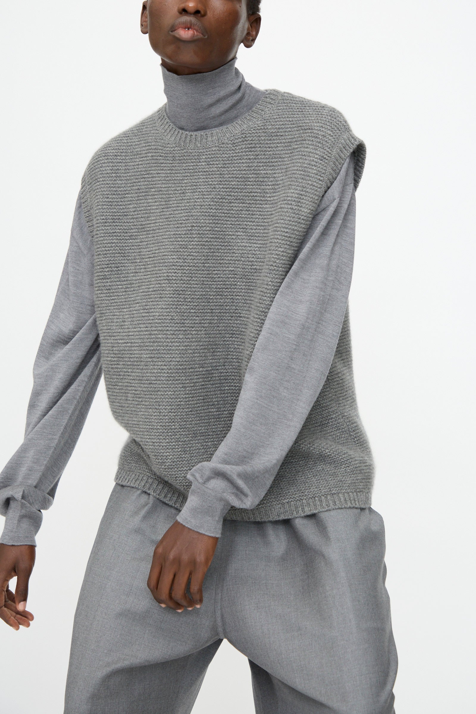 Model wearing the Sofie D'Hoore Cashmere Matta Sweater in Mid Grey Melange under a sleeveless knit vest with loose grey pants, posed against a plain white background.
