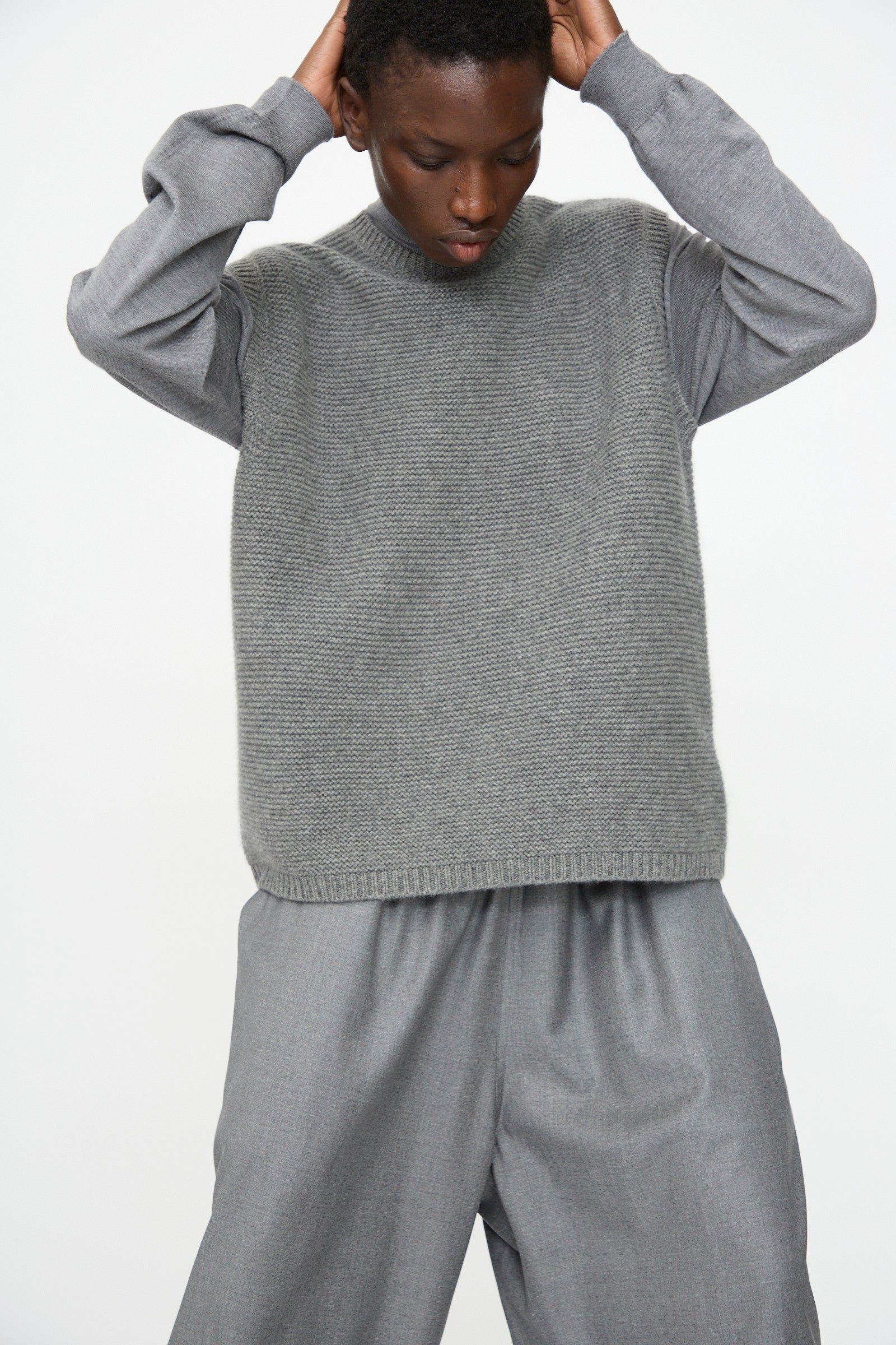 A person wears the Sofie D'Hoore Cashmere Matta Sweater in Mid Grey Melange over a long-sleeved shirt and wide-leg gray pants, standing against a plain light background with hands on their head.