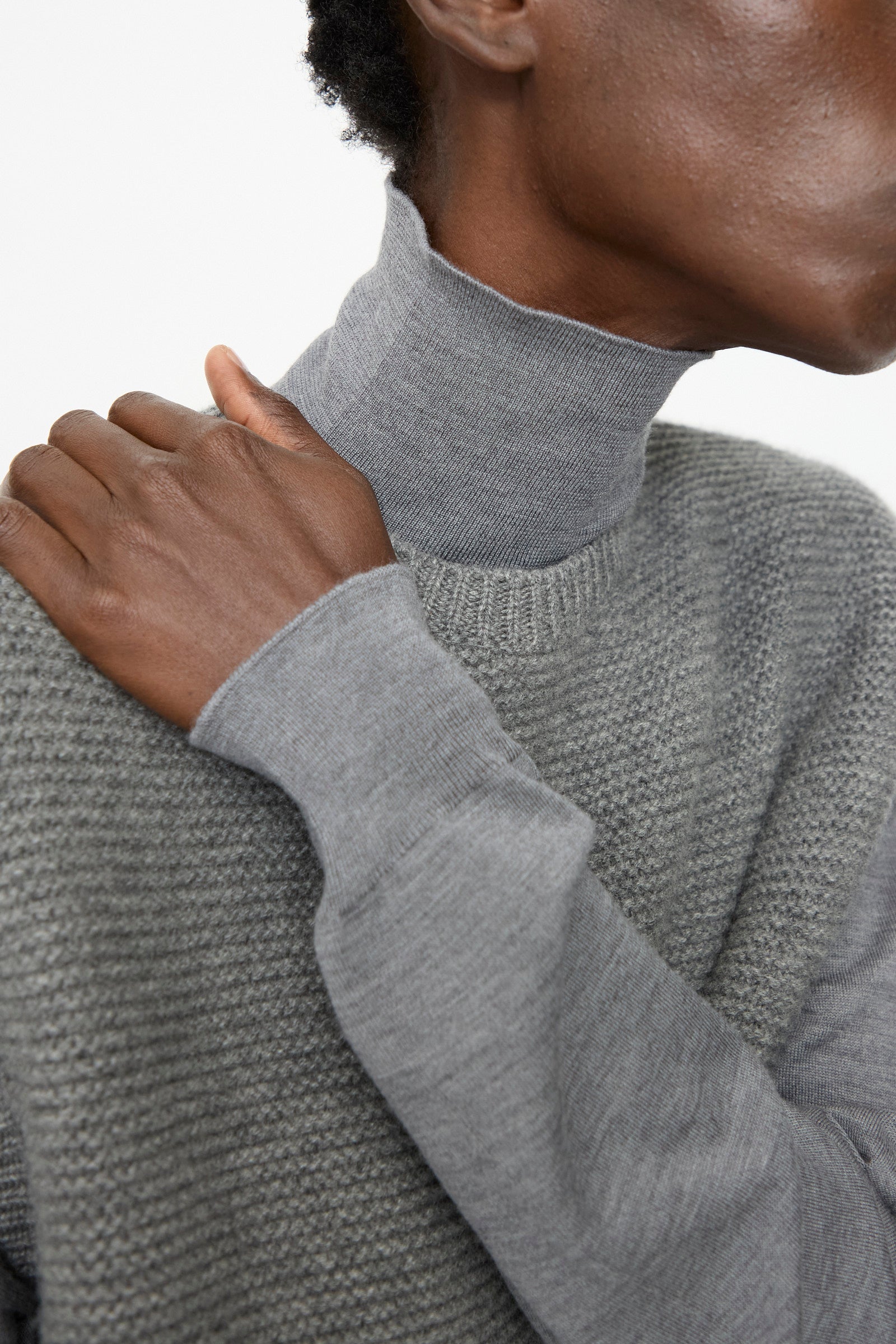 A person wears the Sofie D'Hoore Cashmere Matta Sweater in Mid Grey Melange over a light gray turtleneck, with one hand resting on their shoulder.