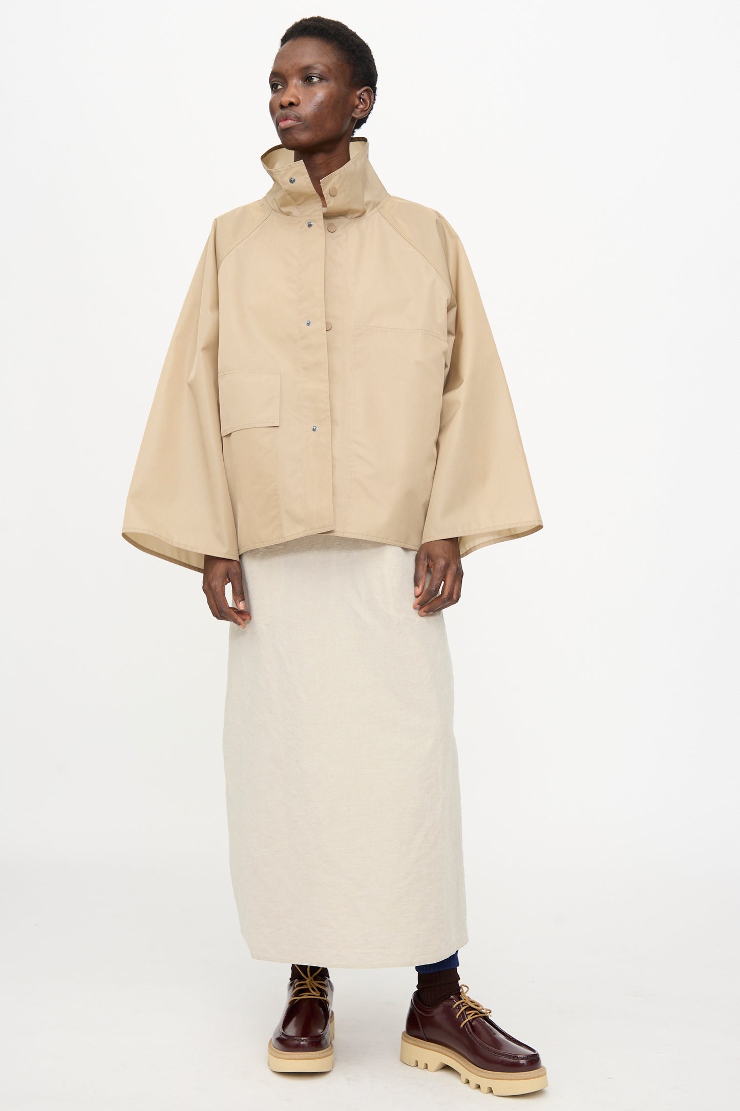 A person wears the Sofie D'Hoore Ceil Jacket in Moonstone—an oversized fit with wide sleeves—paired with a long cream skirt and brown lace-up boots, standing against a plain white background.