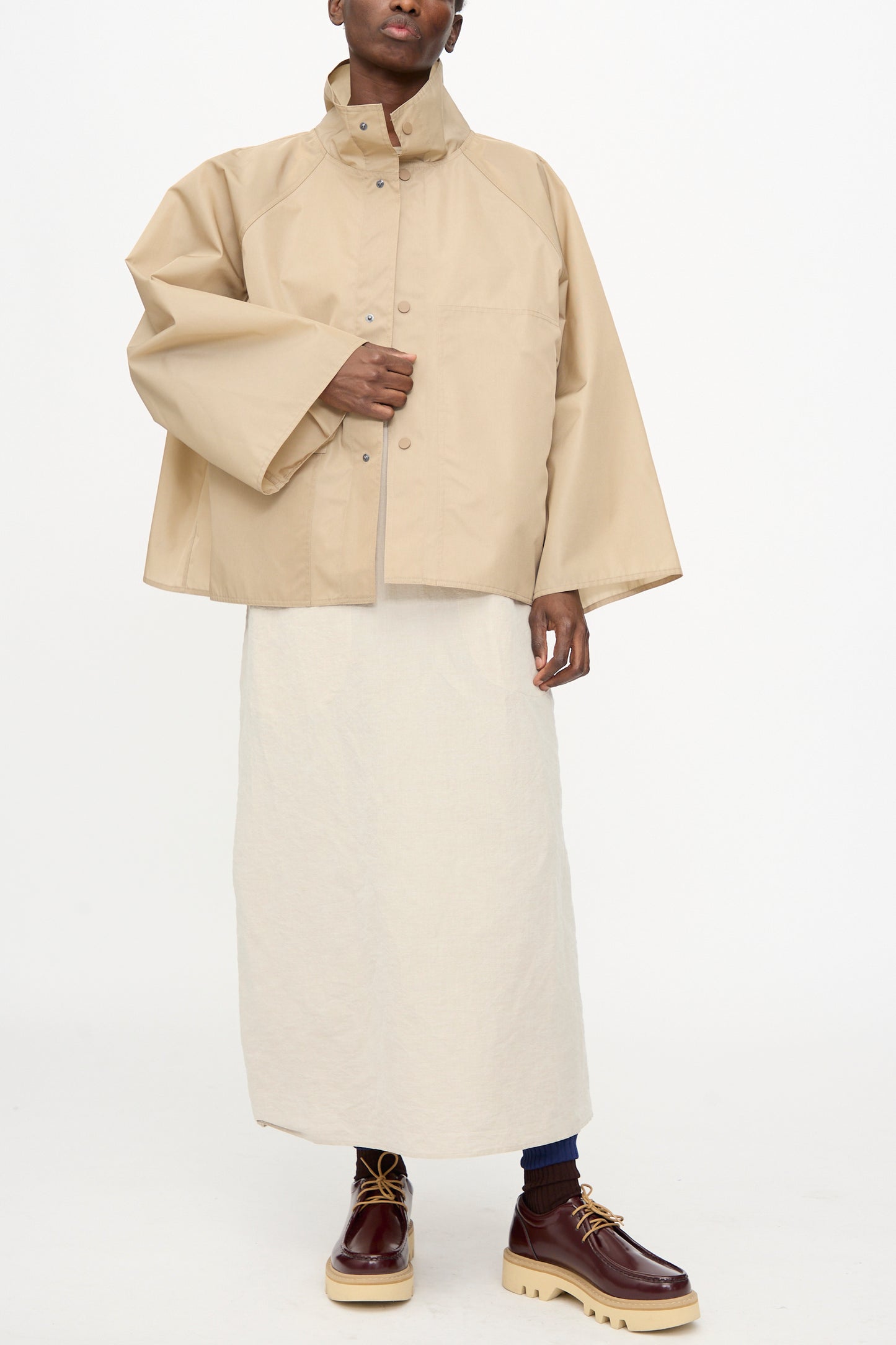 A person wearing the Sofie D'Hoore Ceil Jacket in Moonstone, styled with an off-white skirt, dark socks, and brown platform shoes, stands against a plain white background.