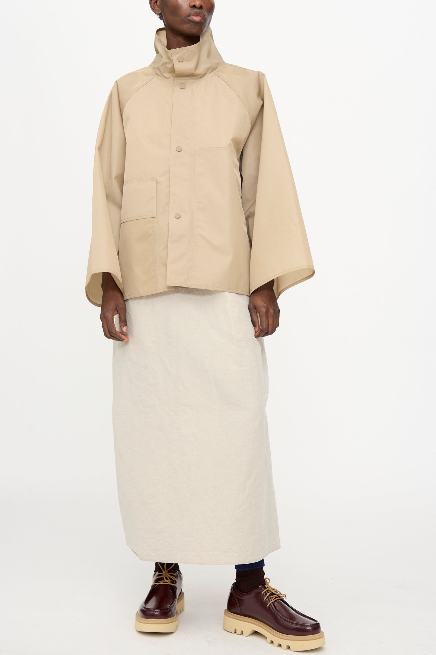 A person wearing the Sofie D'Hoore Ceil Jacket in Moonstone, paired with a cream long skirt and brown lace-up shoes, stands against a plain white background.