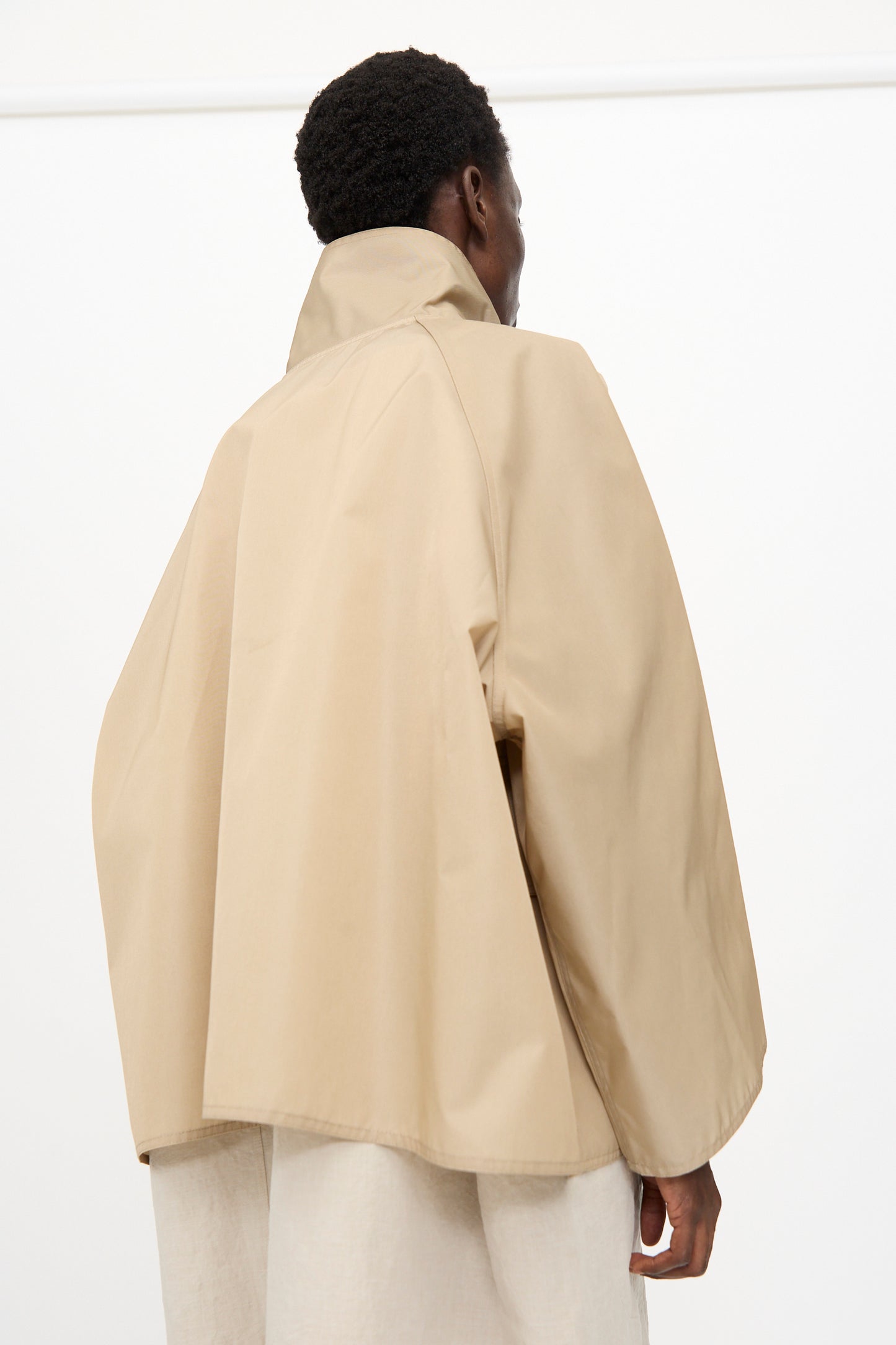 Person faces away, wearing the Ceil Jacket in Moonstone by Sofie D'Hoore—a light beige, oversized taffeta canvas jacket with a high collar. Plain white background.