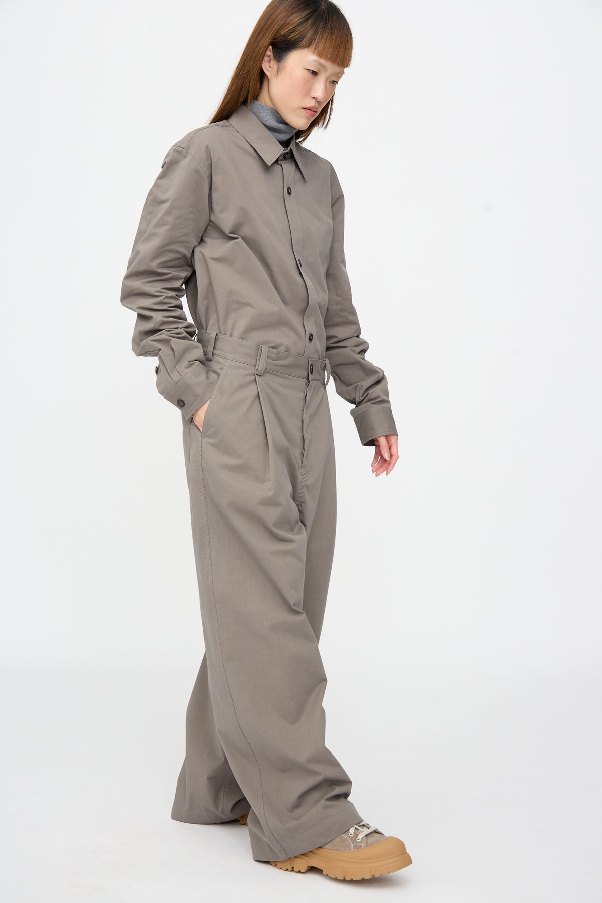 A person wearing a long-sleeved, button-up shirt and Sofie D’Hoore Compact Winter Cotton Pasadena Pant in Turtle, paired with tan-soled sneakers.