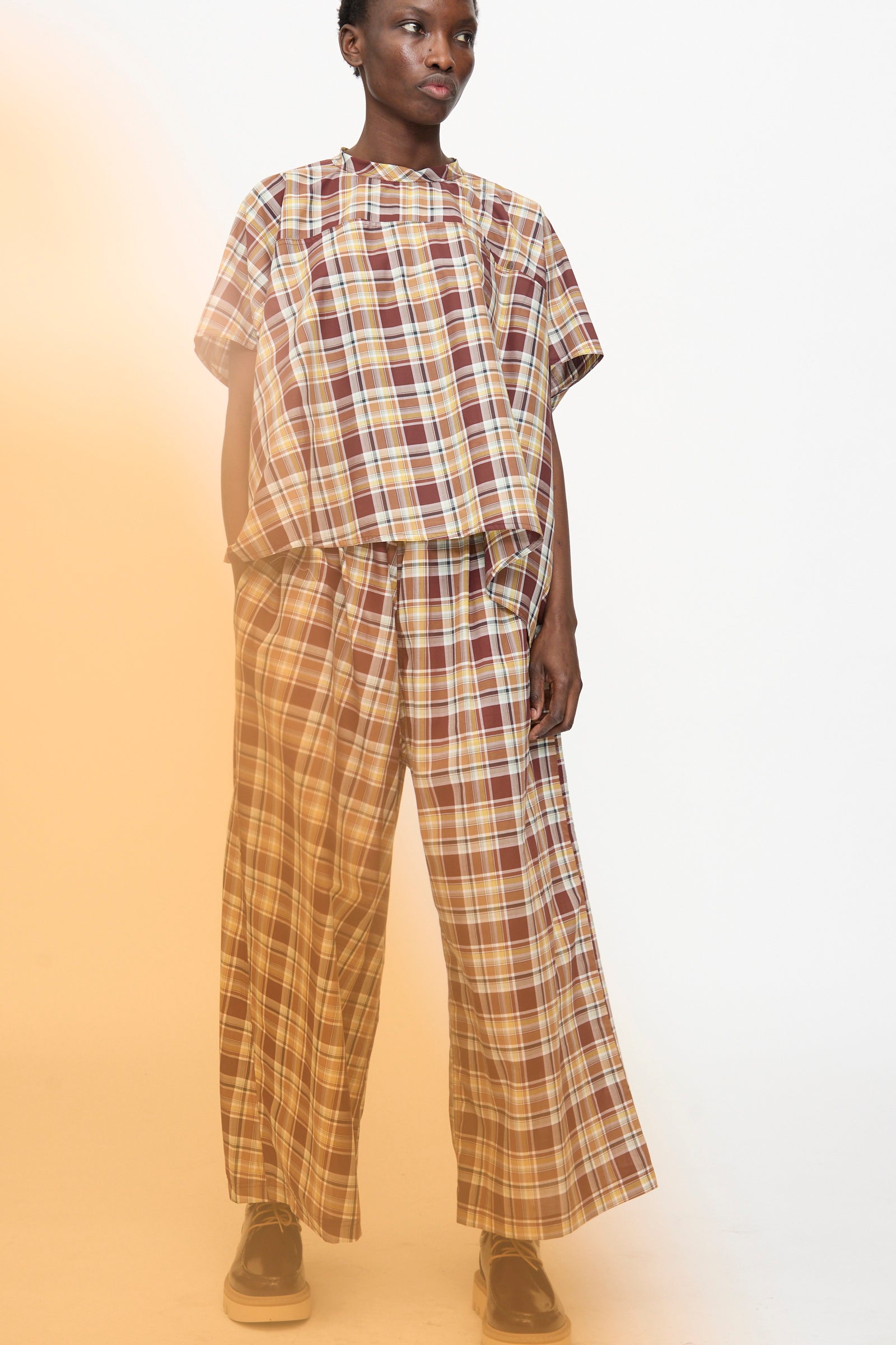 A person wears the Sofie D'Hoore Cotton Batiste Check Basker Shirt in Toffee, styled loosely with matching wide-leg plaid pants and black shoes, standing against a plain background.