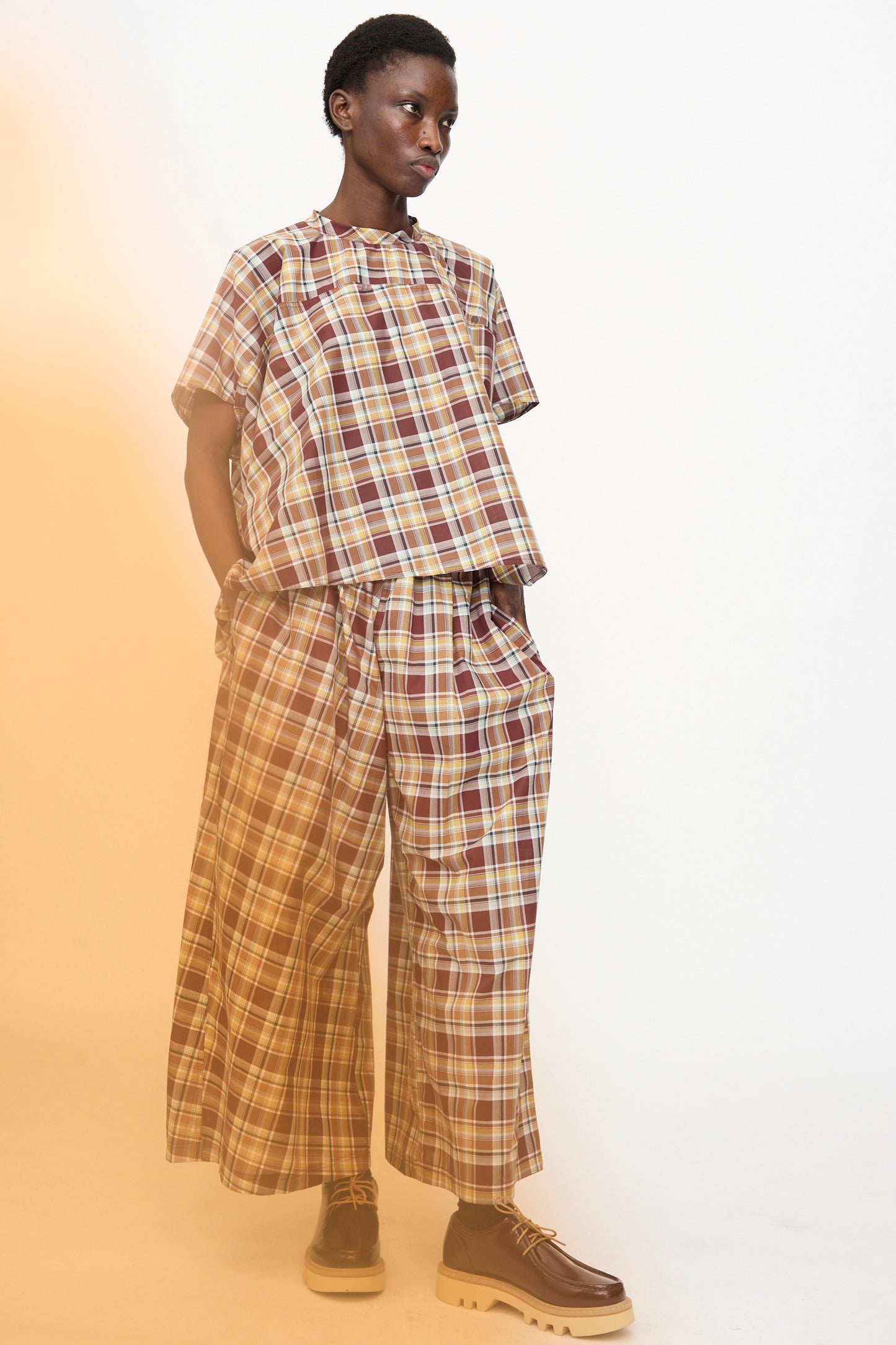 A person stands against a plain background wearing the Sofie D'Hoore Cotton Batiste Check Basker Shirt in Toffee, styled boxy with short sleeves, paired with matching wide-leg pants with pockets and brown platform shoes.