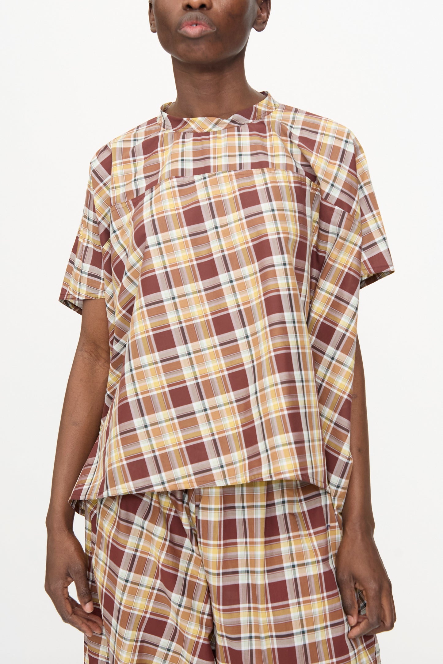A person wears the Sofie D'Hoore Cotton Batiste Check Basker Shirt in Toffee—short sleeves, boxy fit, brown/yellow/white plaid—paired with matching shorts, standing against a plain white background.