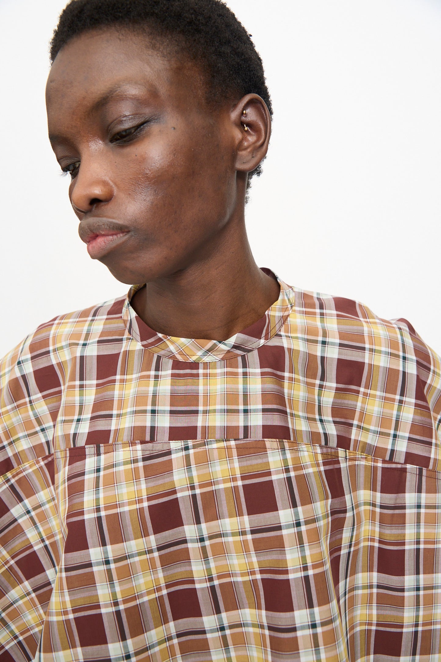 A person with short hair wears the Sofie D'Hoore Cotton Batiste Check Basker Shirt in Toffee—a loose, high-neck, boxy-fit plaid shirt in brown, yellow, and white—against a plain white background.