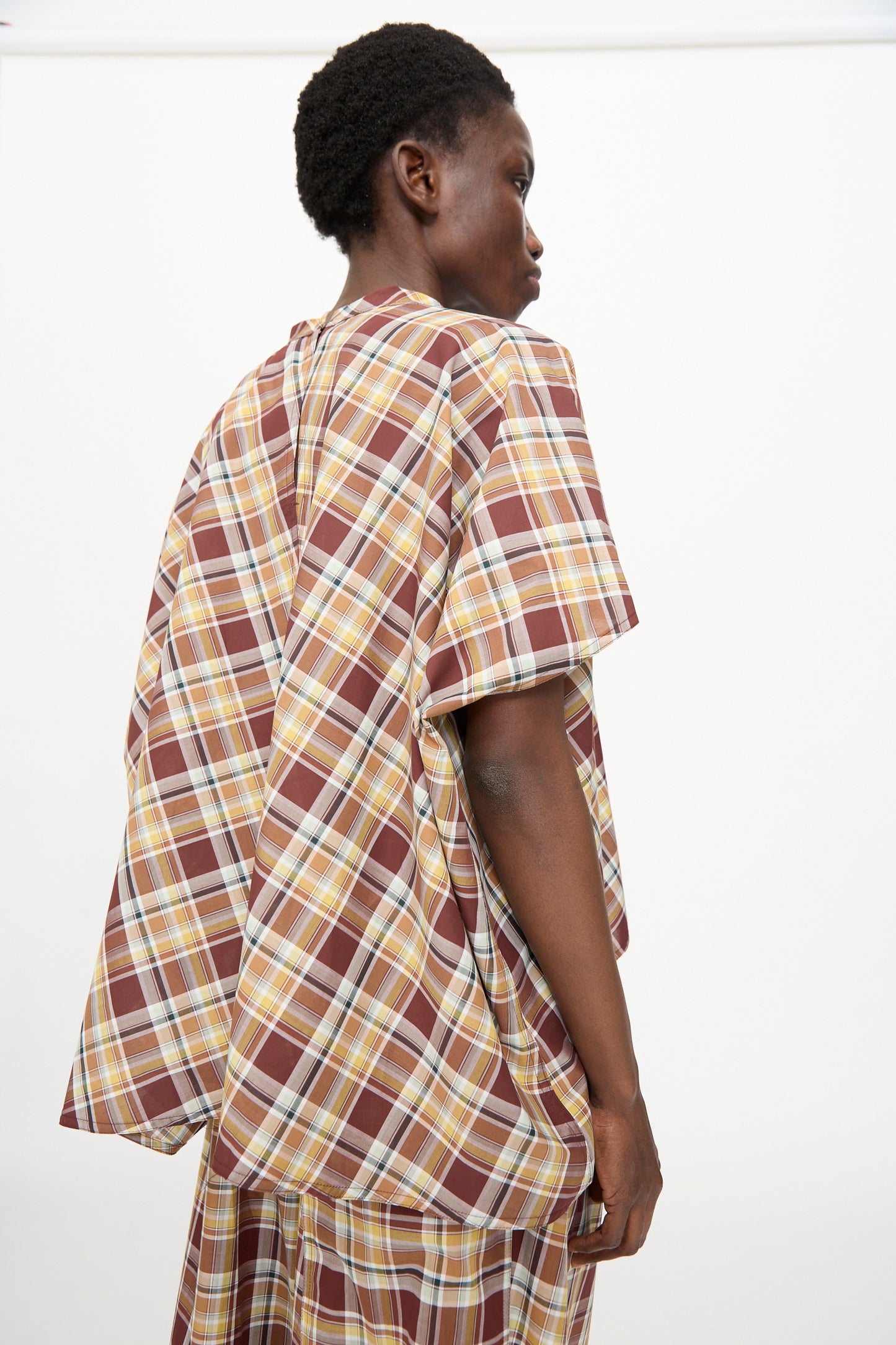 A person is seen from the side against a plain white background, wearing the Sofie D'Hoore Cotton Batiste Check Basker Shirt in Toffee—a loose-fitting, boxy short sleeve shirt styled with a matching skirt.