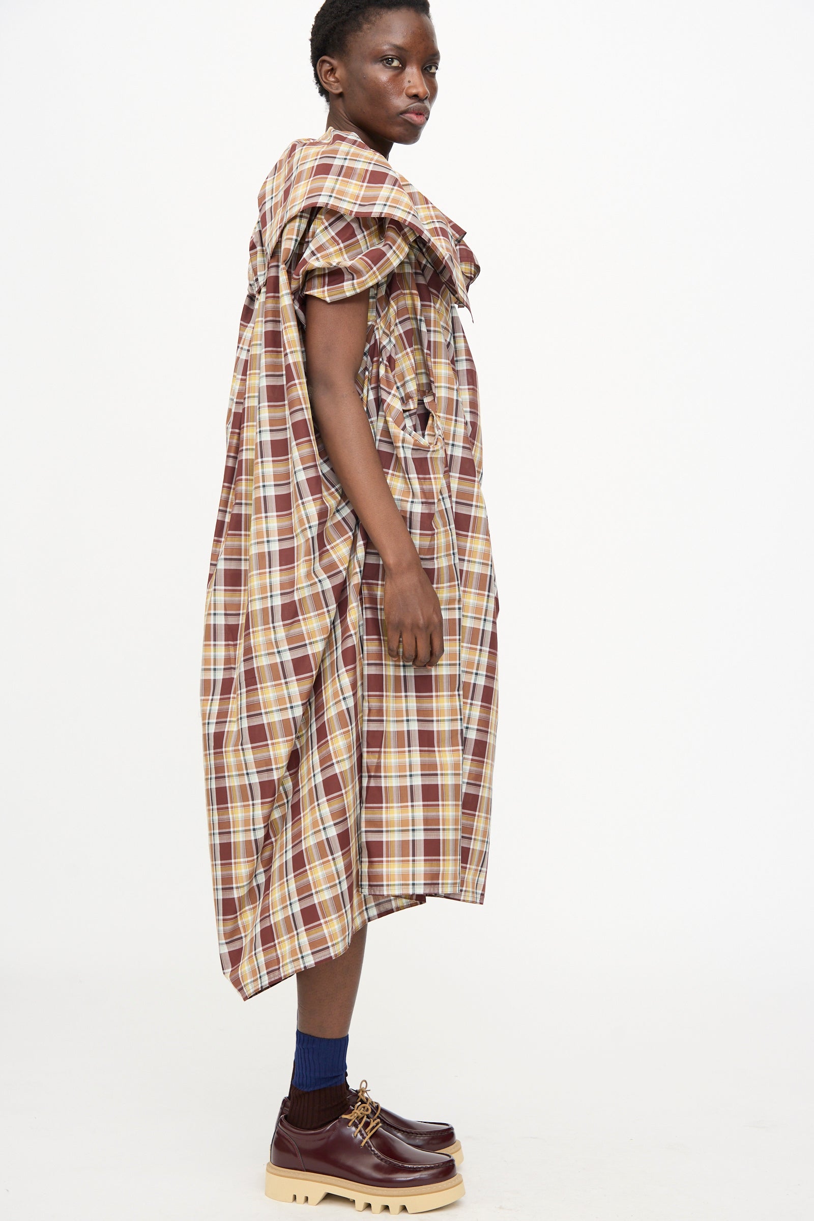 A person stands sideways against a white background wearing the Sofie D'Hoore Cotton Batiste Check Dolabella Dress in Toffee, styled oversized with short sleeves and ruffle detail, paired with blue socks and brown platform shoes.