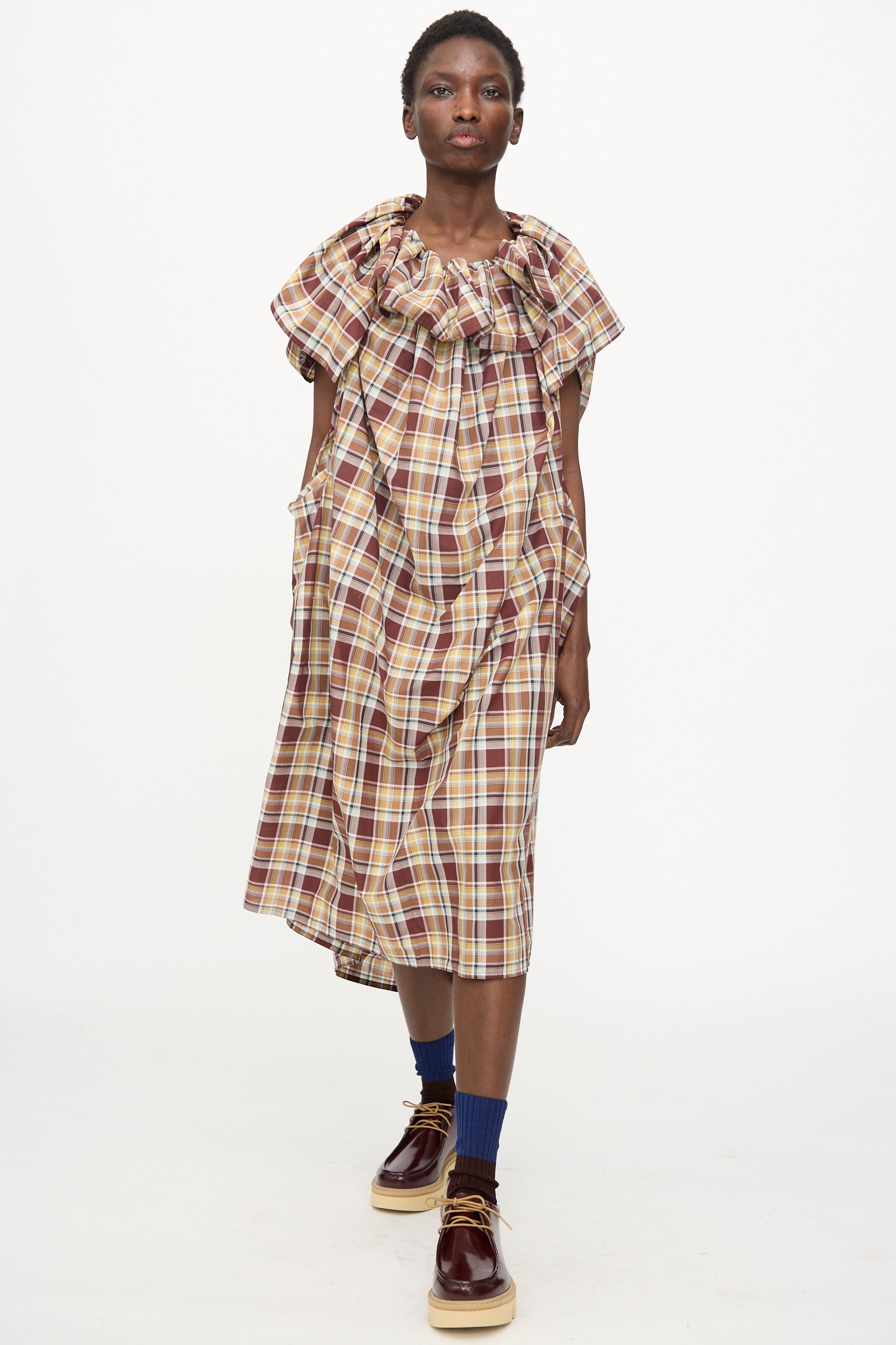 A person wearing the Sofie D'Hoore Cotton Batiste Check Dolabella Dress in Toffee, paired with blue socks and brown shoes, stands against a plain white background.