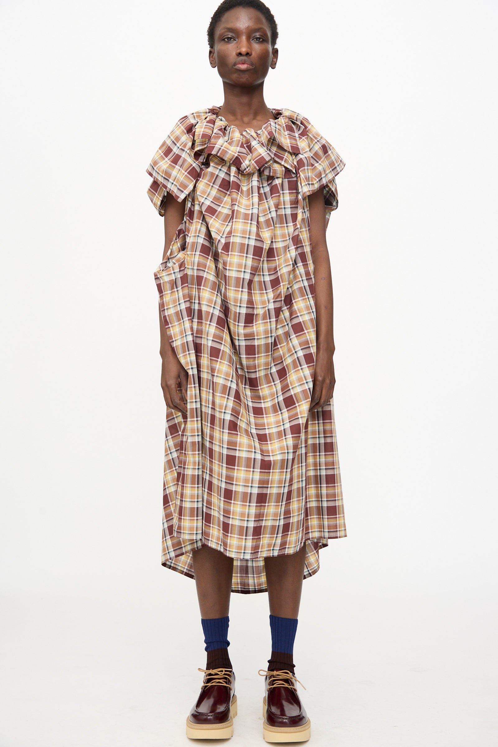 A person faces forward wearing the Sofie D'Hoore Cotton Batiste Check Dolabella Dress in Toffee—an oversized, ruffled plaid midi dress—paired with blue socks and brown shoes against a plain white background.