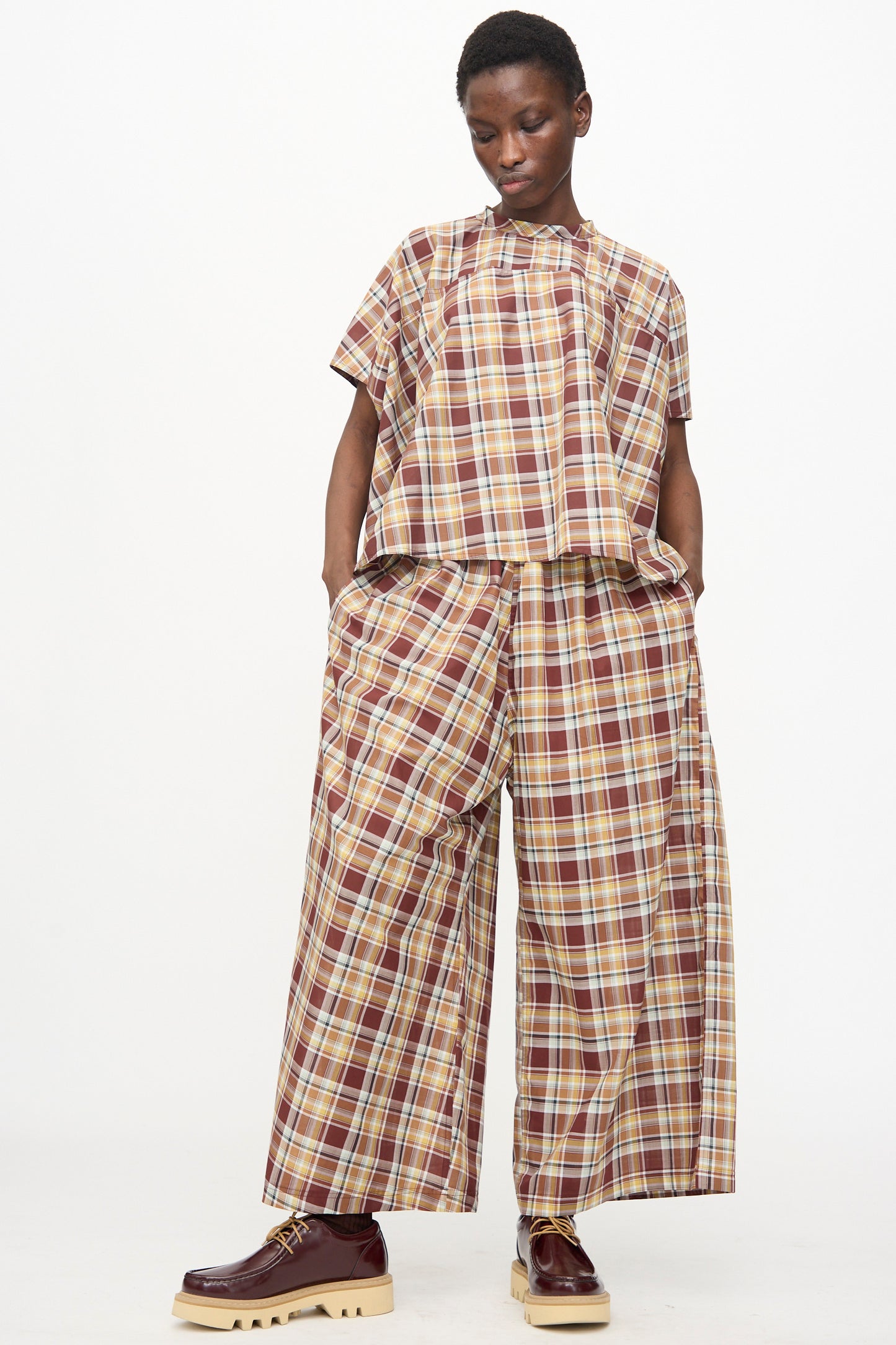 A person stands against a white background wearing Sofie D'Hoore's Cotton Batiste Check Parana Pant in Toffee, styled with a loose short-sleeve top and maroon platform shoes.