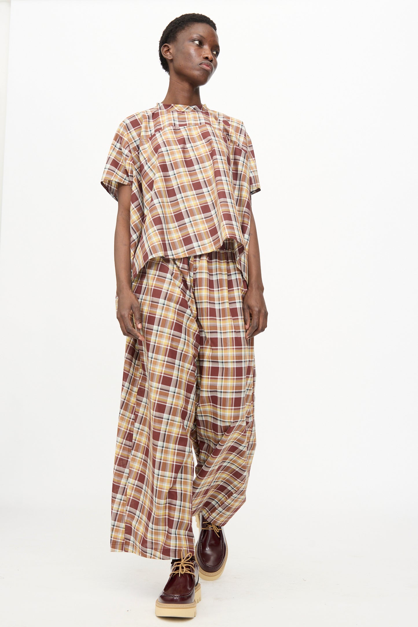 Model in Sofie D'Hoore's Cotton Batiste Check Parana Pant in Toffee, styled with a loose short-sleeve plaid top and maroon shoes, poses against a plain white background.