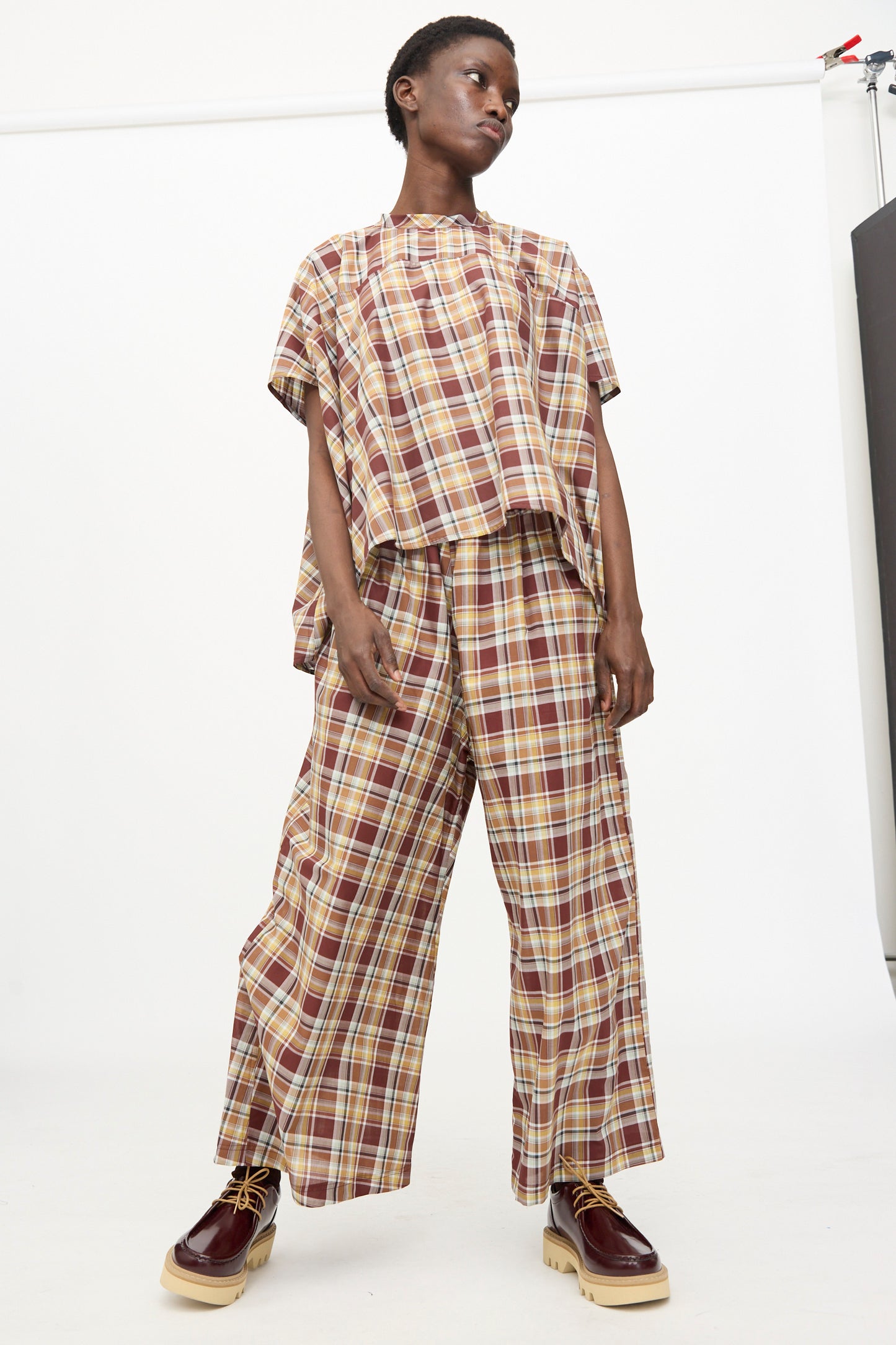 A person stands against a white background wearing Sofie D'Hoore's Cotton Batiste Check Parana Pant in Toffee, styled with a loose plaid top and maroon platform shoes.