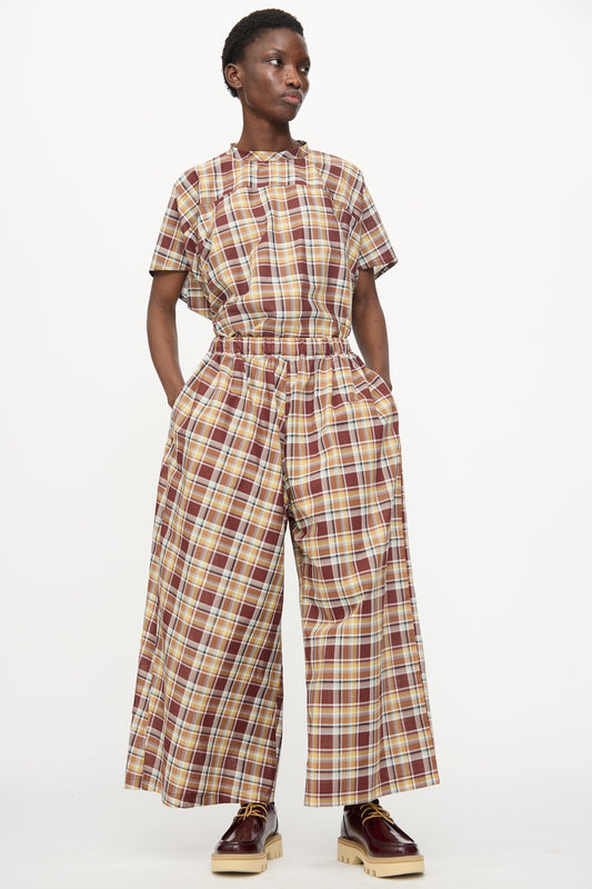 A person stands against a plain background wearing Sofie D'Hoore's Cotton Batiste Check Parana Pant in Toffee—oversized checkered pants paired with a matching plaid short-sleeve top, hands in pockets, and brown platform shoes.