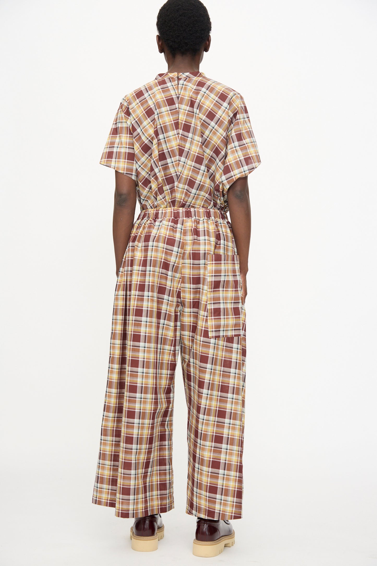 Person facing away from the camera, wearing Sofie D'Hoore's Cotton Batiste Check Parana Pant in Toffee—a brown, yellow, and white plaid wide-leg style—paired with light-colored shoes.