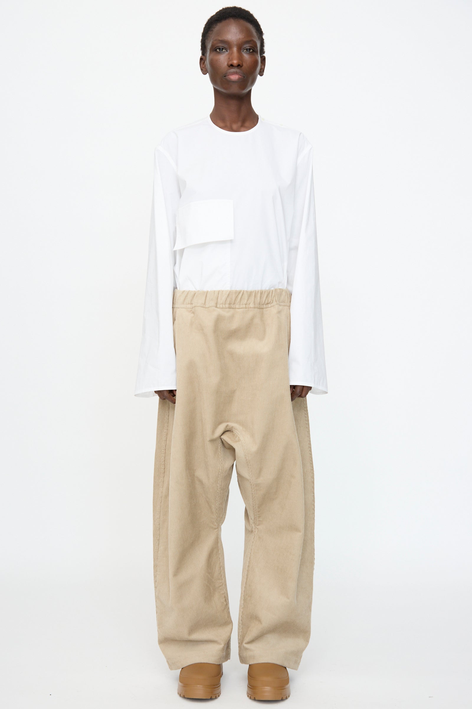 A person stands against a plain white background wearing the Sofie D'Hoore Cotton Corduroy Plof Pant in Cashew, styled with a white long-sleeve shirt and matching tan platform shoes.
