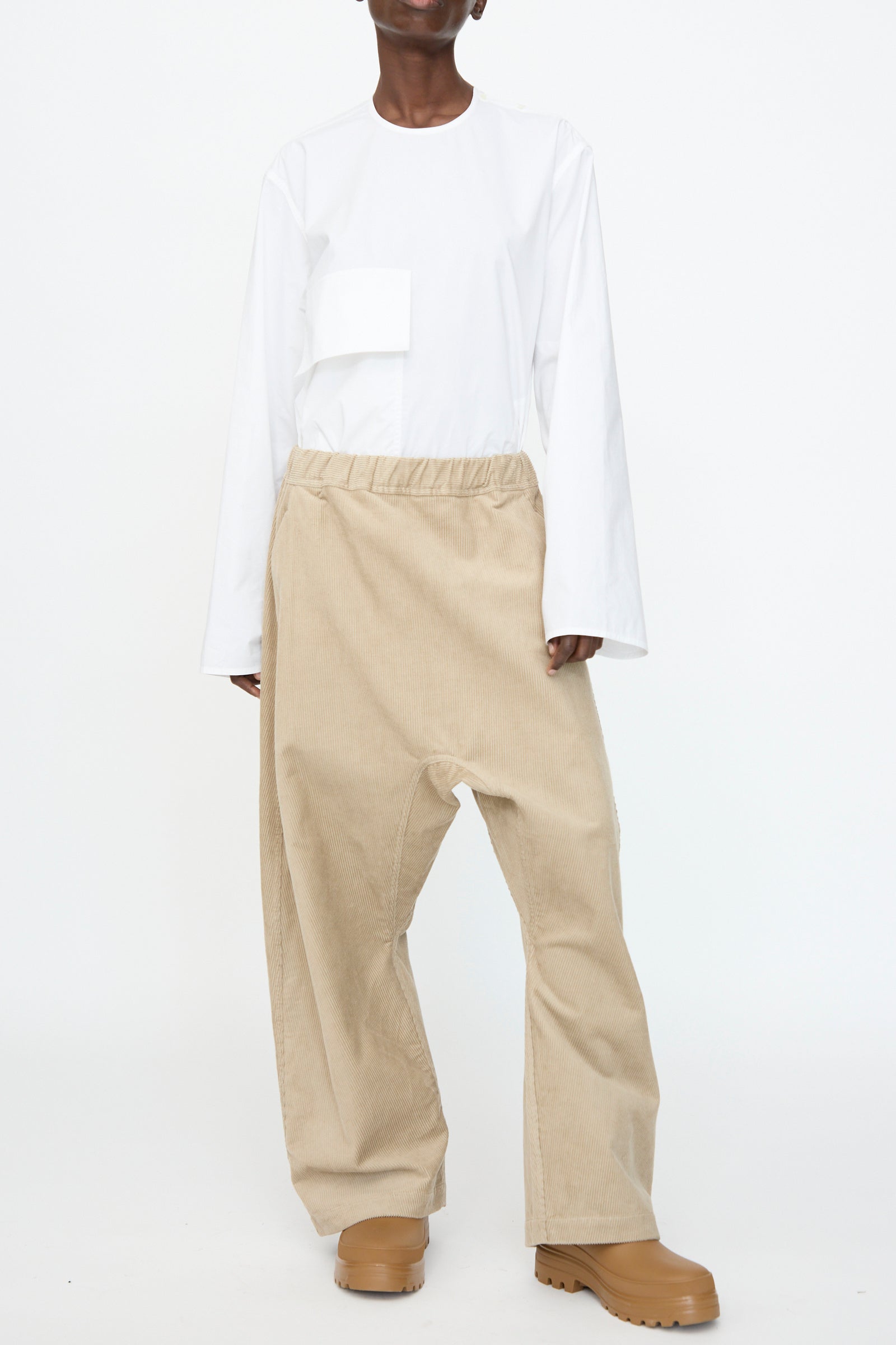 A person stands against a white background wearing a loose white long-sleeve shirt with a front pocket, Sofie D'Hoore's Cotton Corduroy Plof Pant in Cashew, and tan shoes.