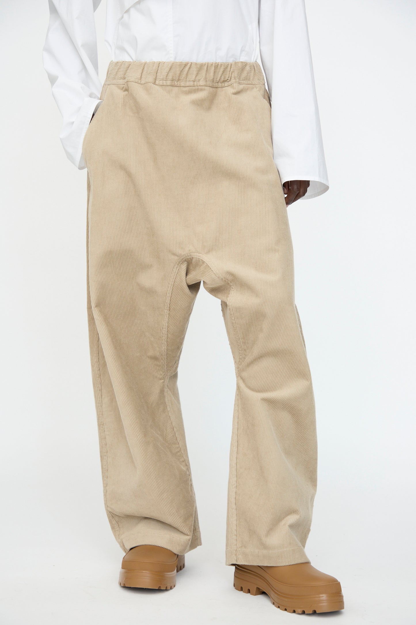 A person wearing Sofie D’Hoore’s Cotton Corduroy Plof Pant in Cashew, paired with a white long-sleeve shirt and light brown chunky shoes, stands with hands in pockets against a plain background.