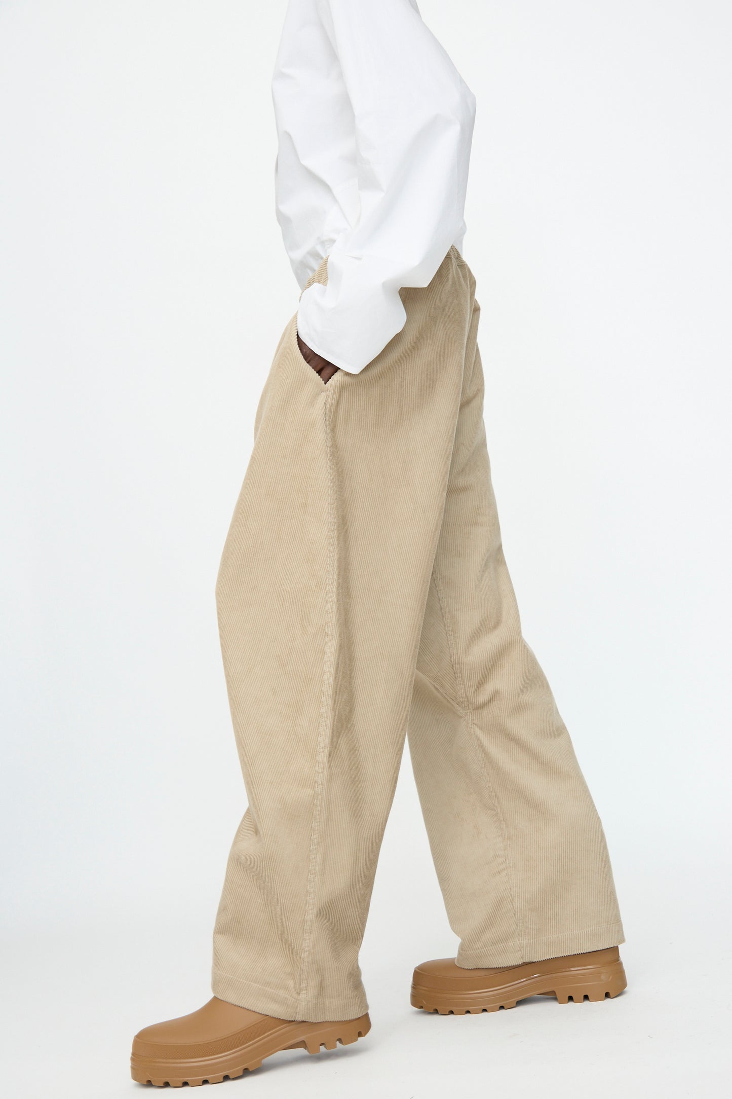 A person wears a white long-sleeve shirt, Sofie D'Hoore Cotton Corduroy Plof Pant in Cashew, and tan rubber boots, photographed walking against a plain white background.