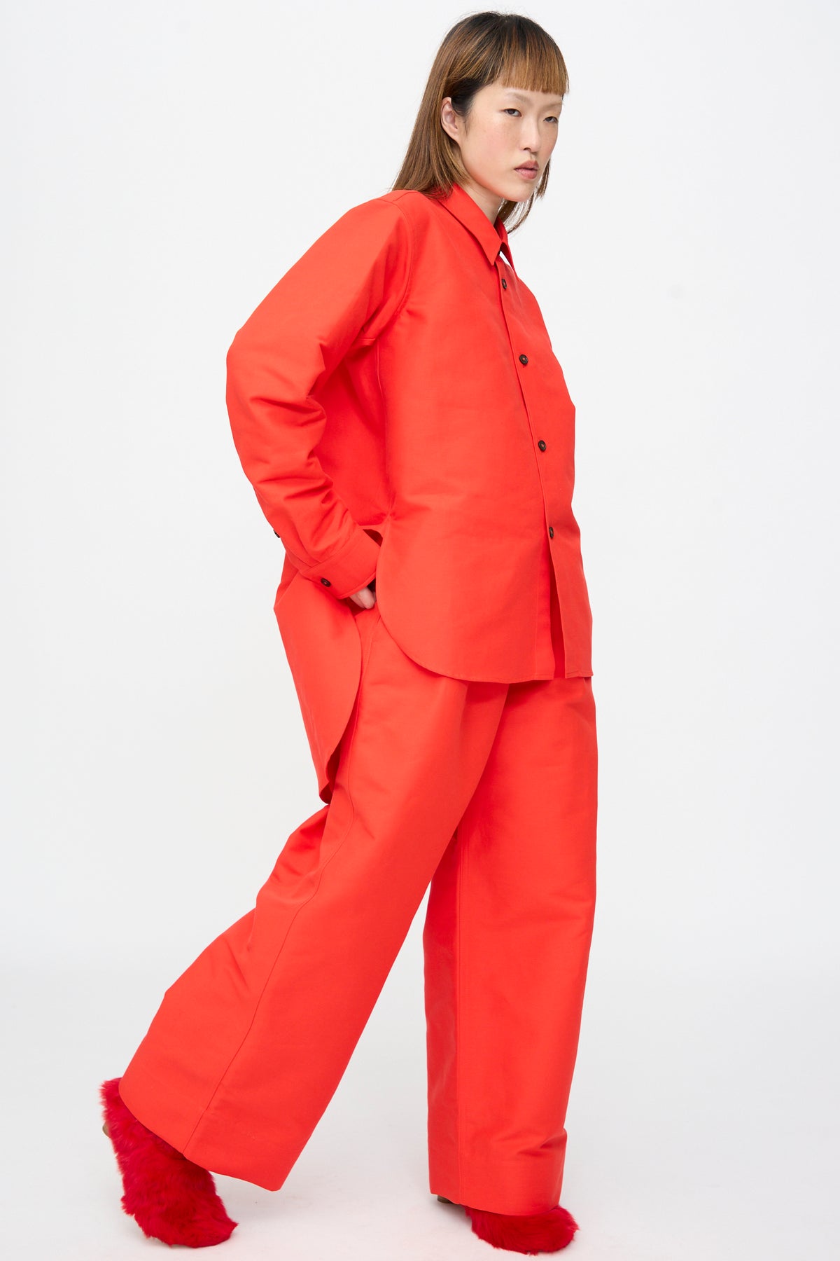 A person wears an oversized Cotton Duchesse Brant Shirt in Fire paired with matching wide-leg trousers, one hand tucked into a pocket.