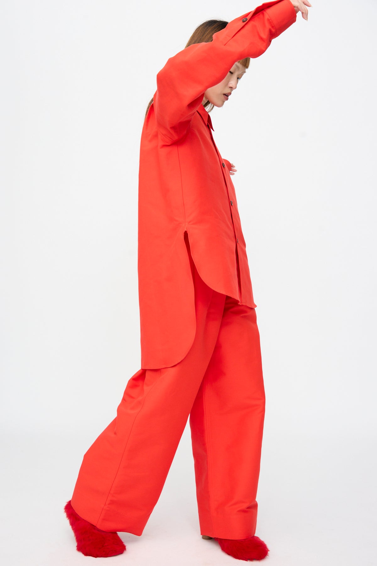 A person wearing a Cotton Duchesse Brant Shirt in Fire, styled as an oversized, long-sleeved red button-down with a hi-low hem, matching wide-leg trousers, and red fuzzy slippers.