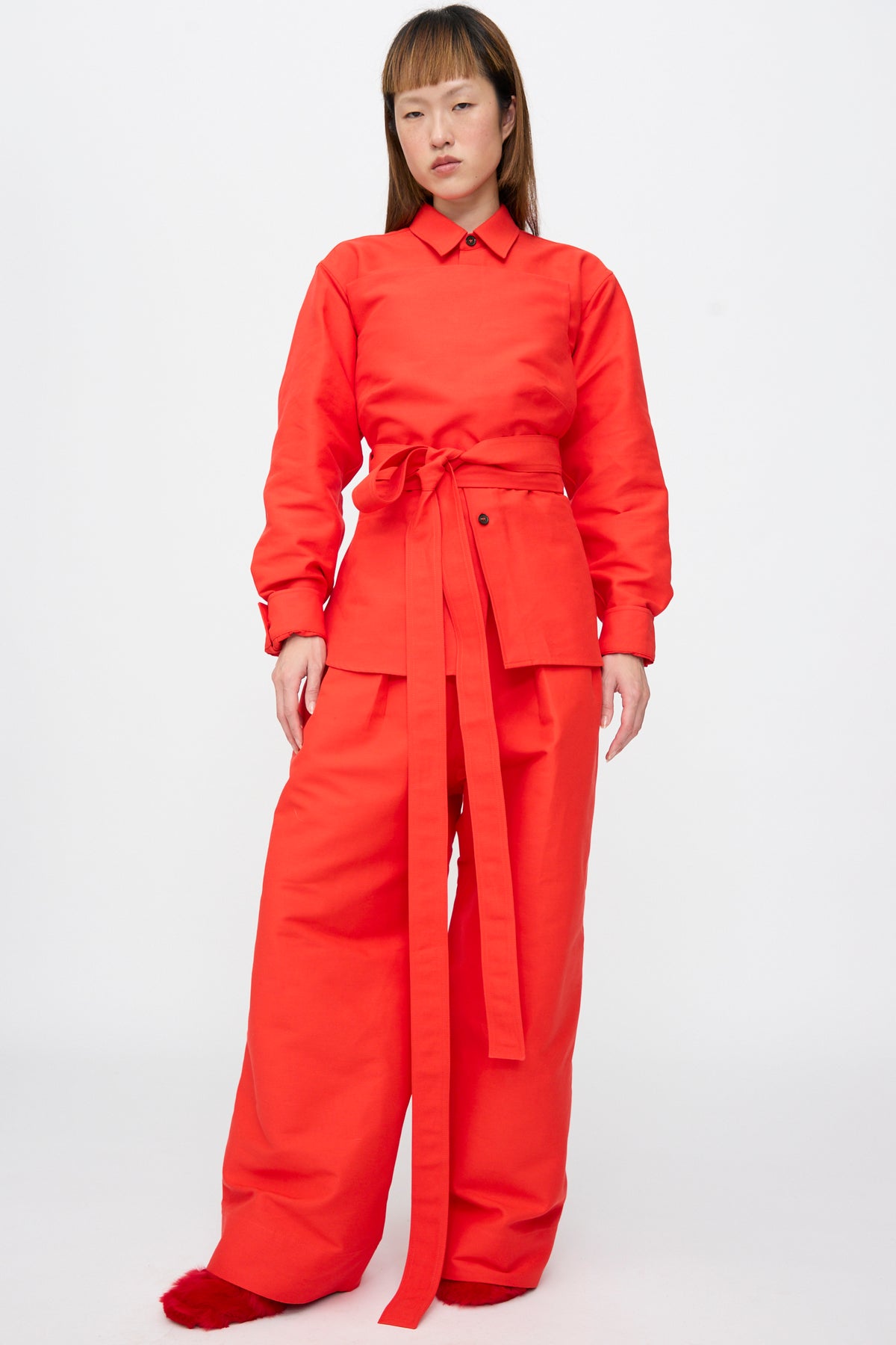 A model wears a bright red Cotton Duchesse Brel Shirt in Fire with a wrap-around waist tie and matching wide-leg trousers.