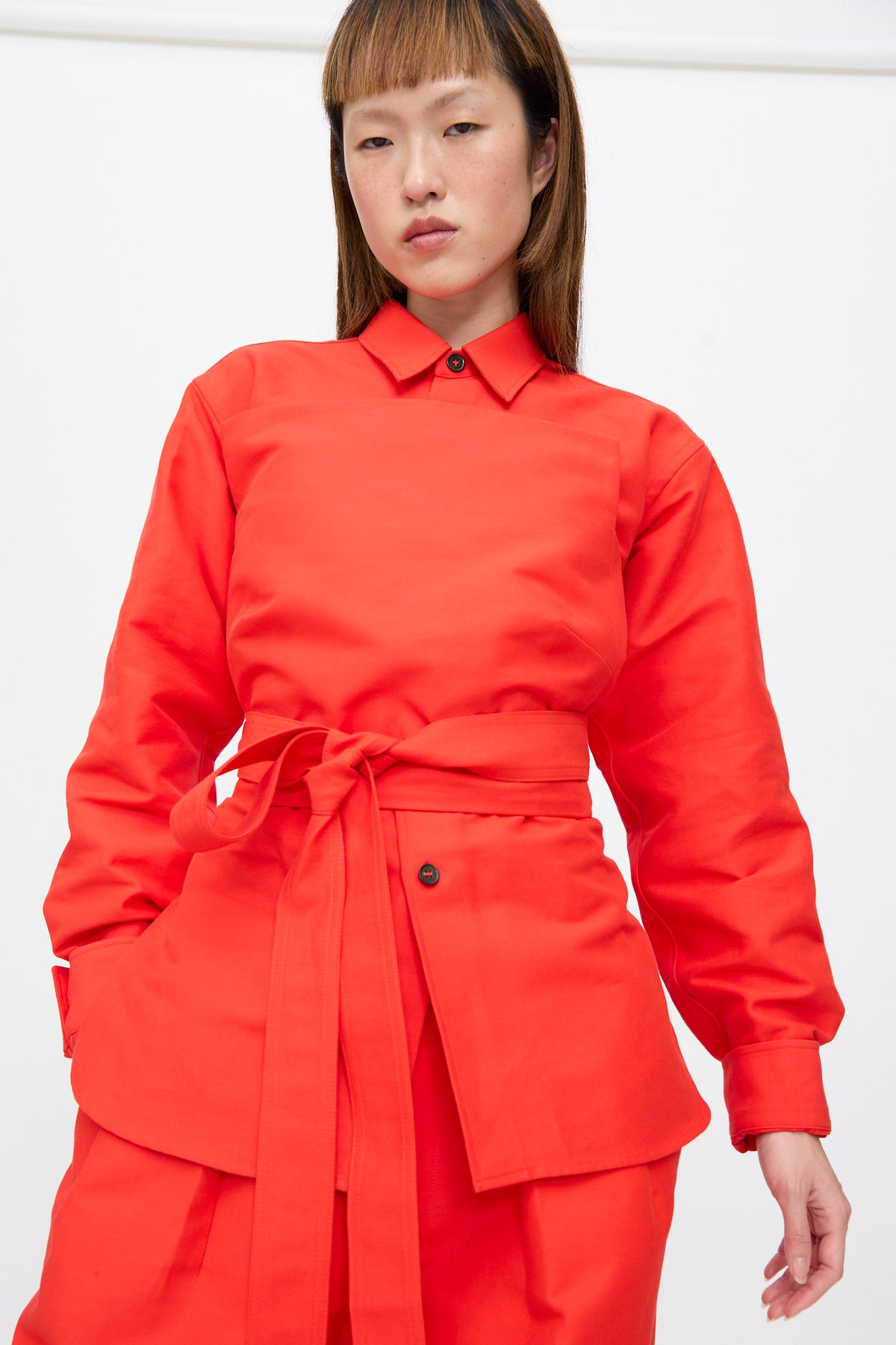 A person wearing a bright red Cotton Duchesse Brel Shirt in Fire, featuring long sleeves, a collar, buttoned placket, and a wrap-around waist tie.