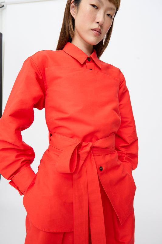 A person wearing a fire-colored Cotton Duchesse Brel Shirt by Sofie D’Hoore, featuring long sleeves, a collar, and a wrap-around tie waist.