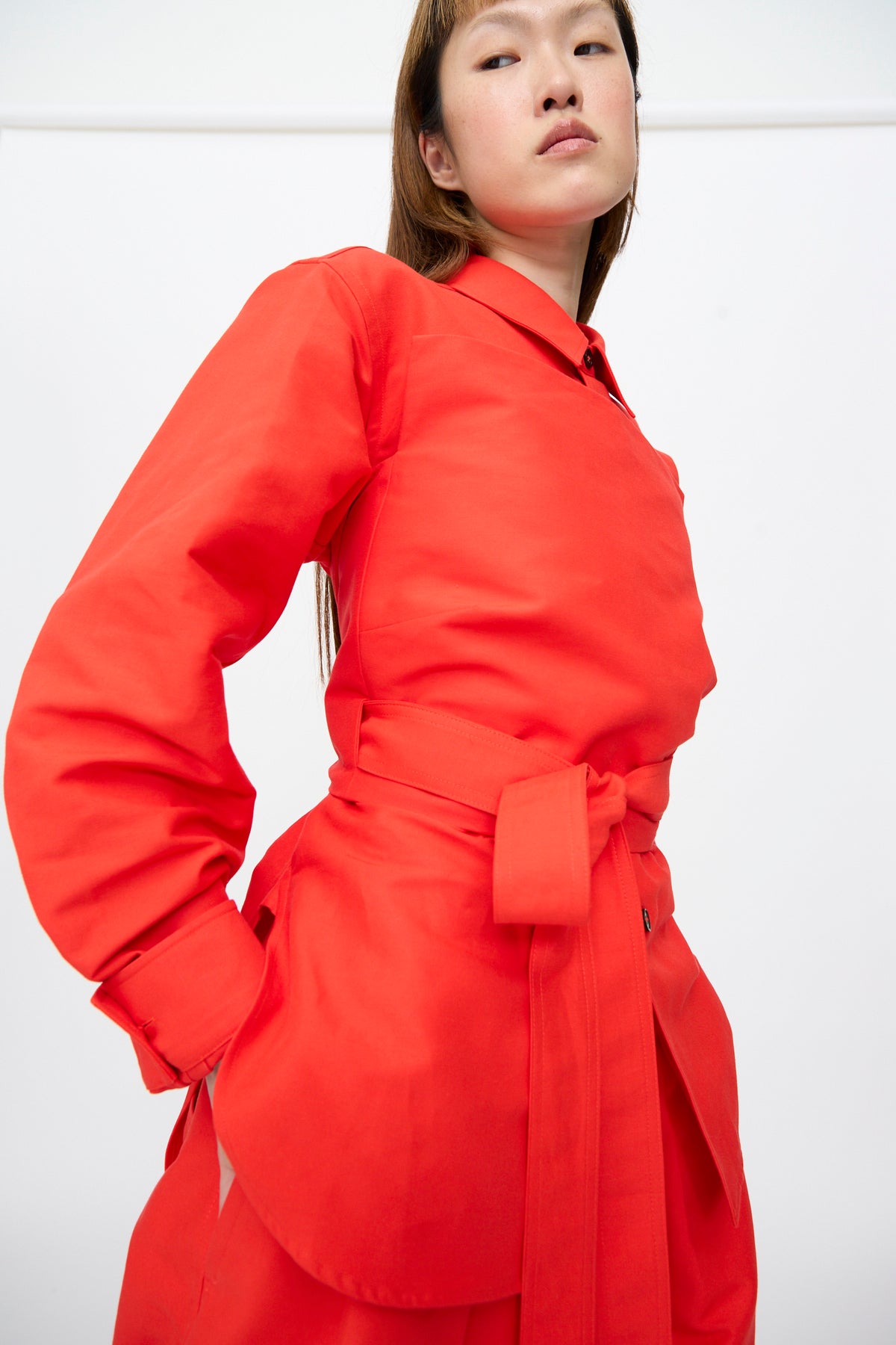 A model wears a bright red Cotton Duchesse Brel Shirt in Fire, featuring long sleeves, a pointed collar, and a wrap-around tie belt at the waist.