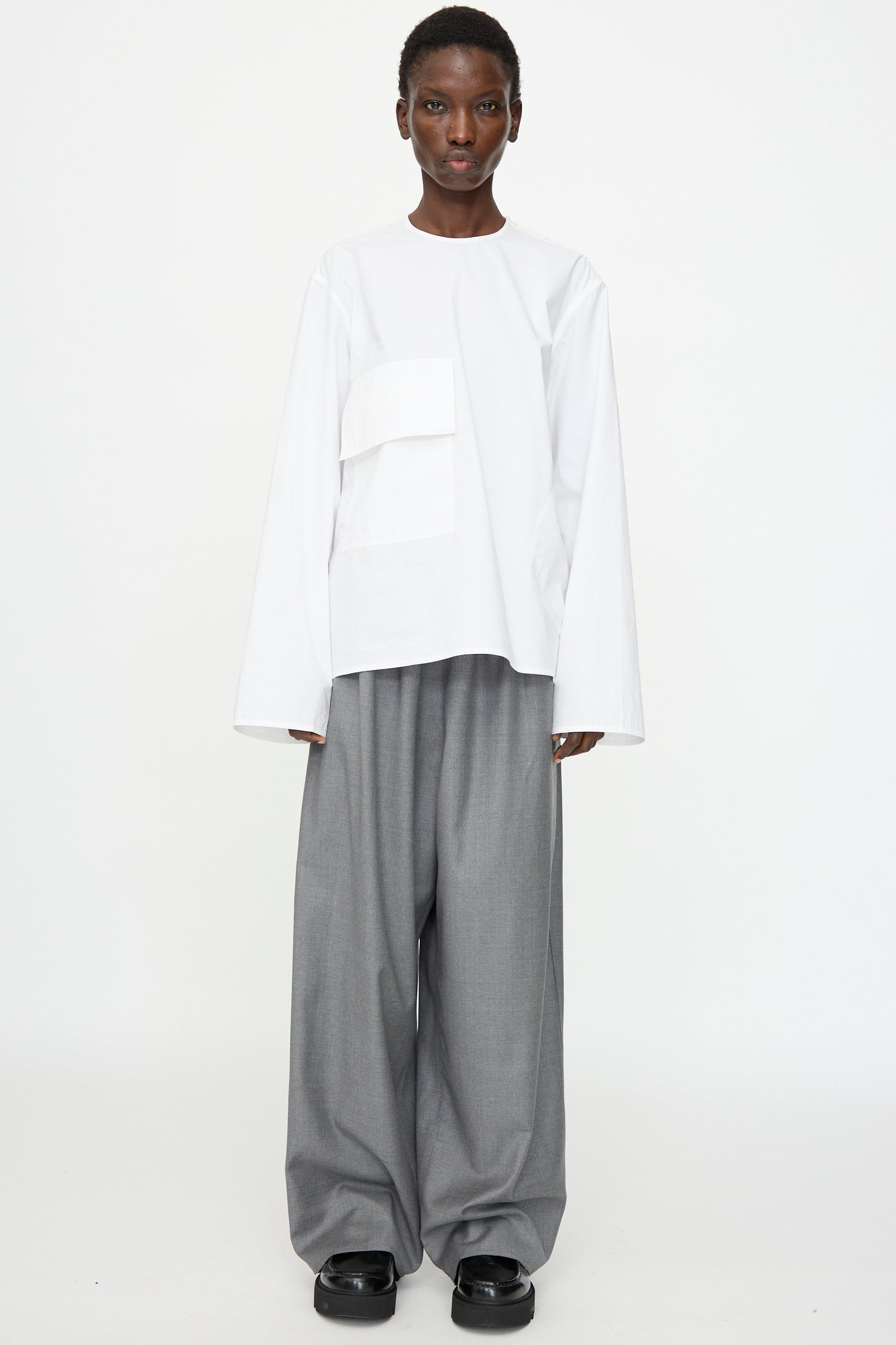 A person wears the Sofie D'Hoore Cotton Poplin Balthus Shirt in White, paired with wide-leg gray trousers and black shoes, standing against a plain white background.