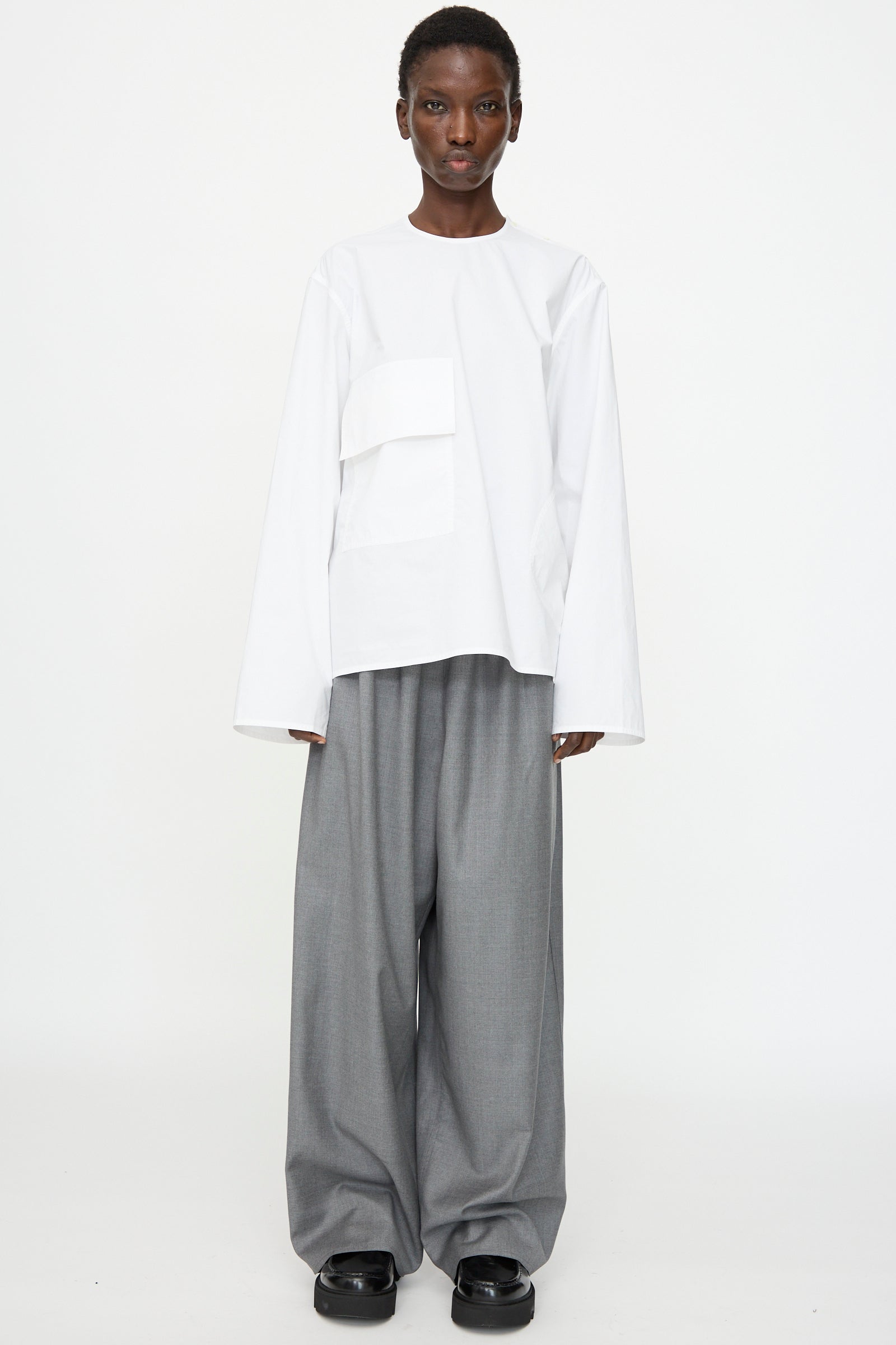 A person wears the Sofie D'Hoore Cotton Poplin Balthus Shirt in White, paired with wide-leg gray trousers and black shoes, standing against a plain white background.