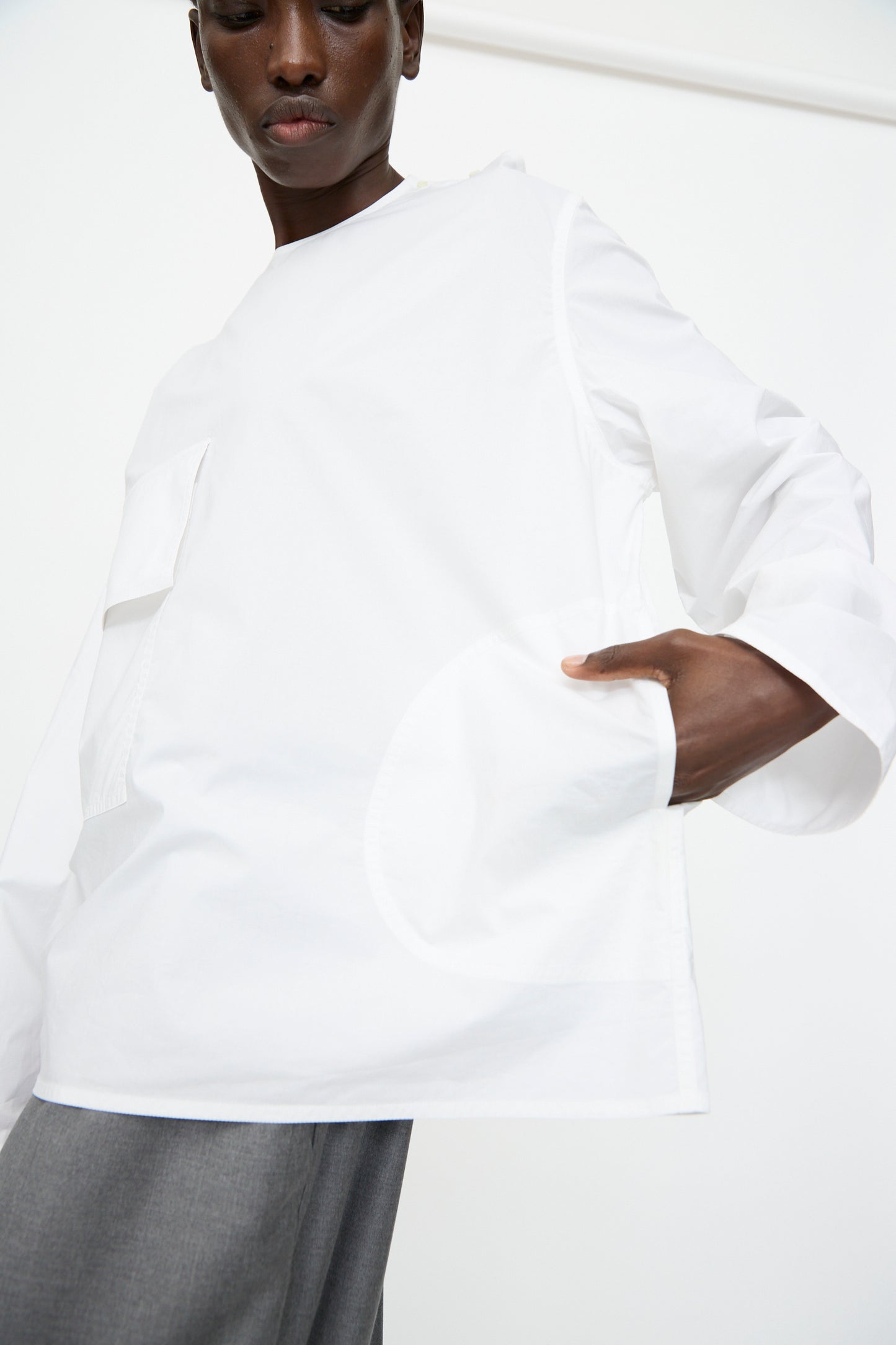 A person wears the Sofie D'Hoore Cotton Poplin Balthus Shirt in White—an oversized long sleeve with a front pocket—paired with gray pants, standing against a plain white background.
